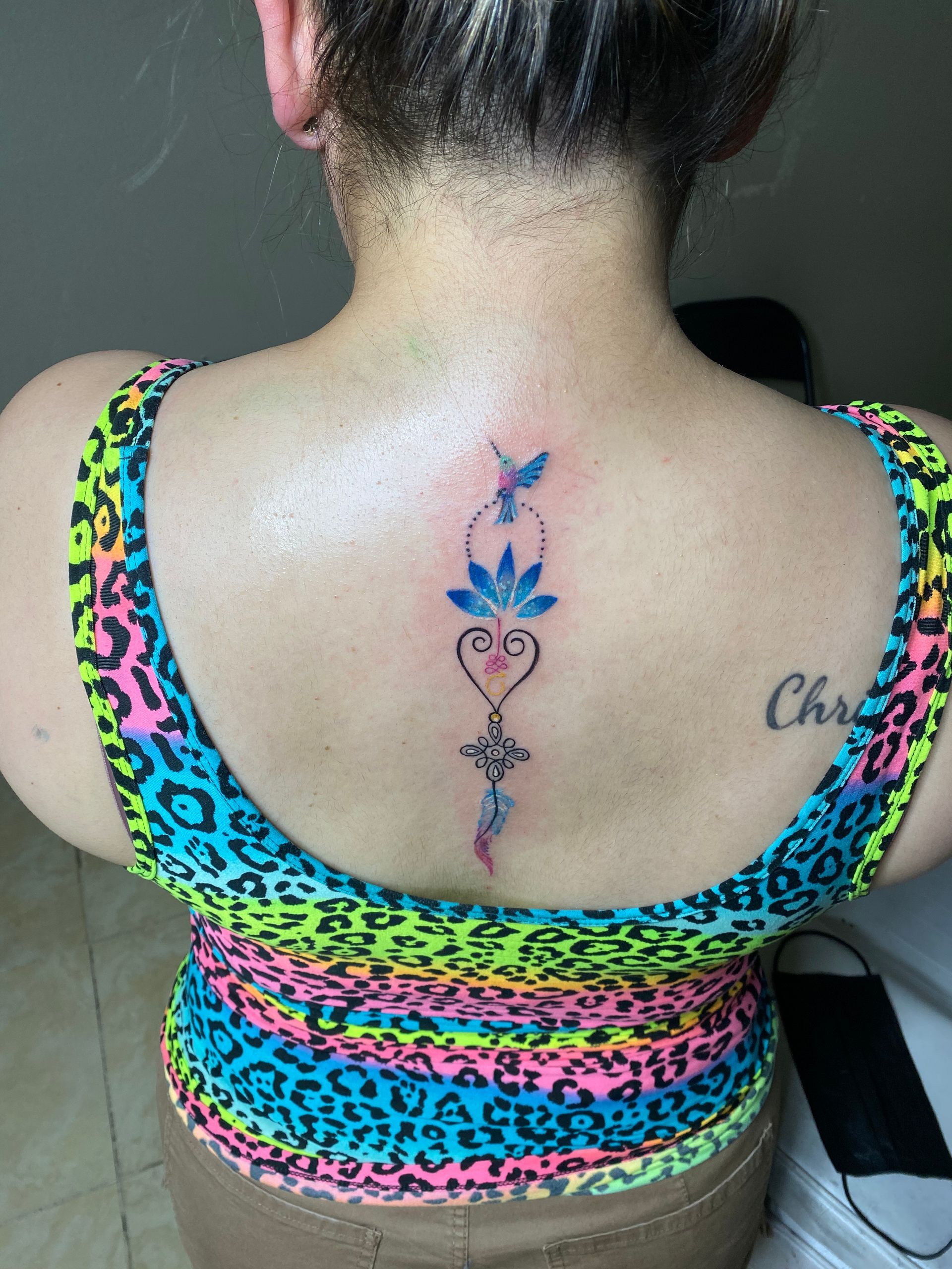 Woman's back with a vertical tattoo of a lotus flower, hummingbird, and heart; colorful top.