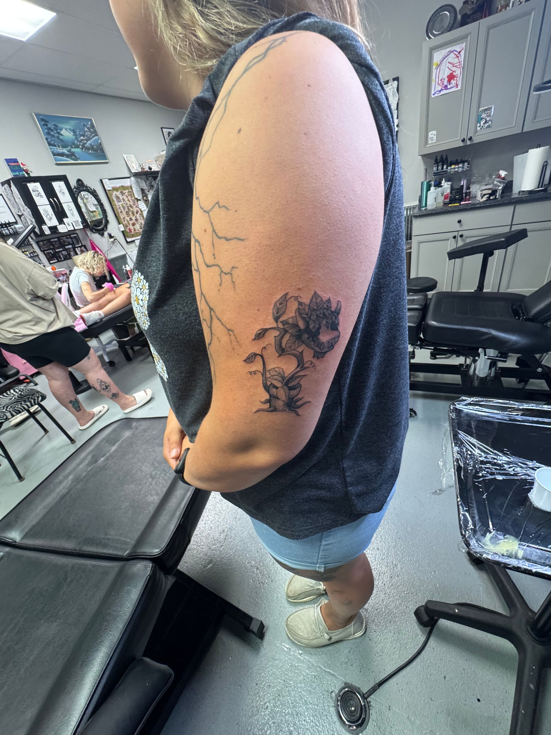 Woman with a floral tattoo on her arm at a tattoo shop, wearing a tank top and shorts.