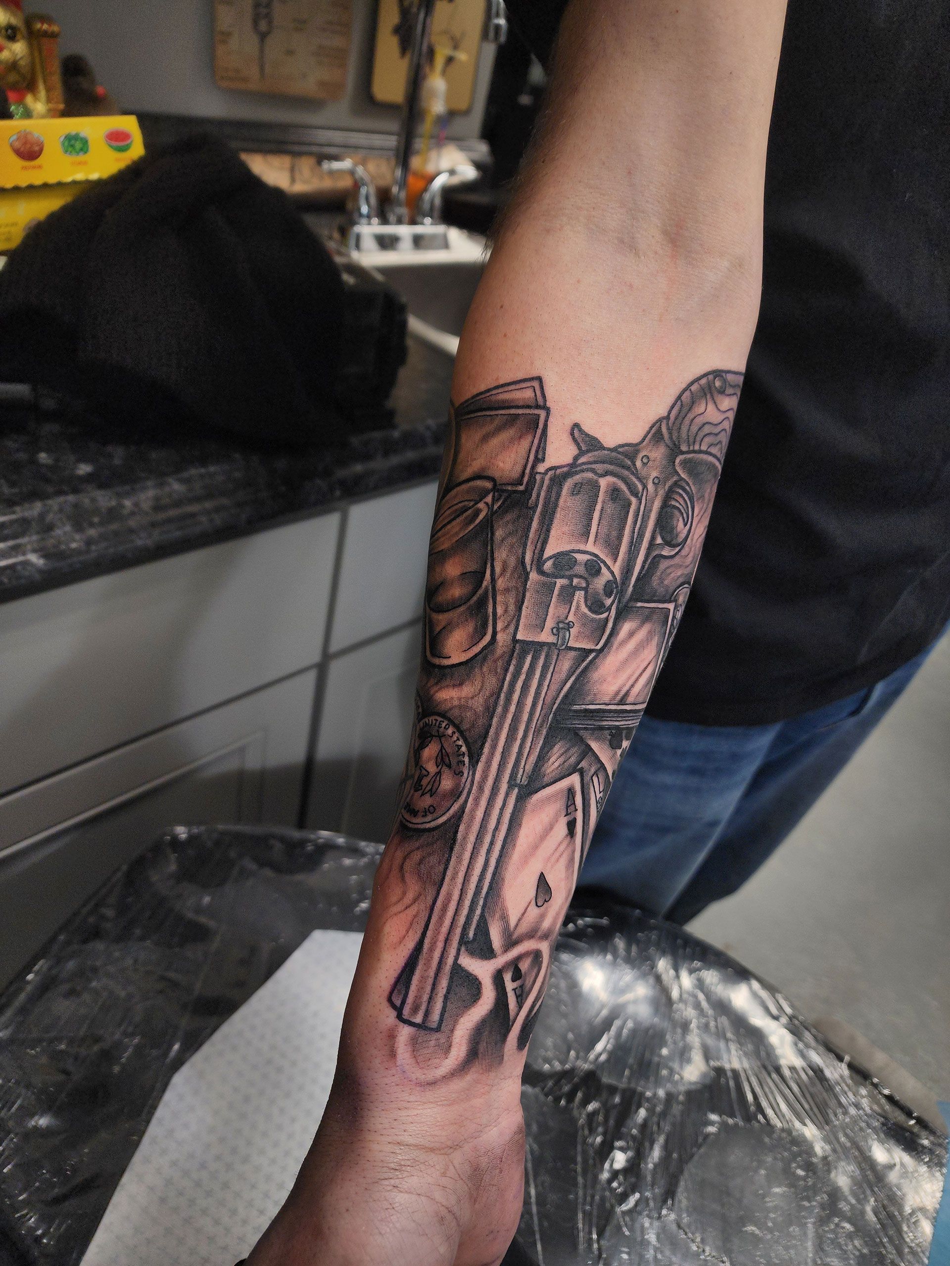 Gun, cards, coins, and drink tattoo on a man's arm