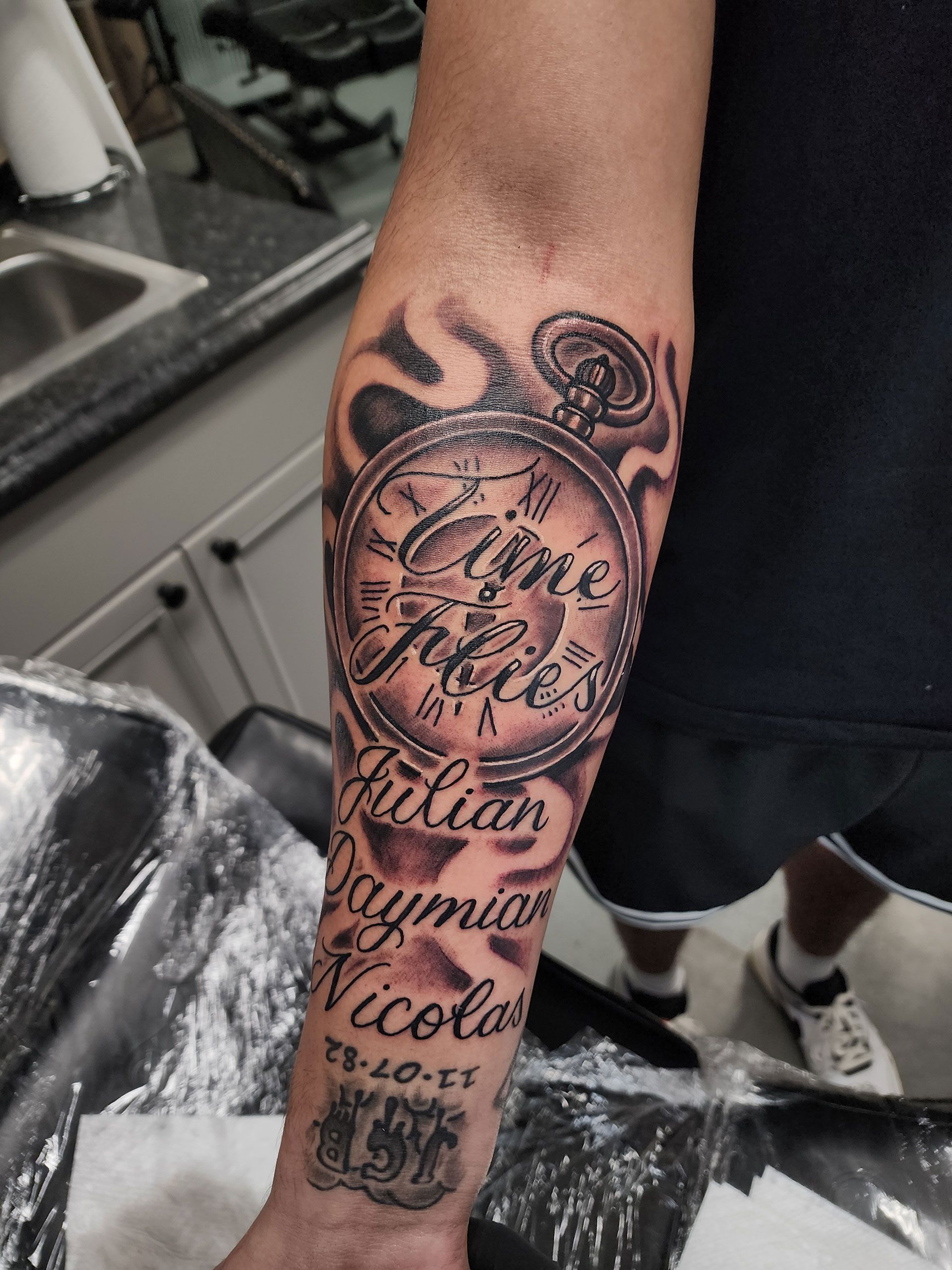 A man has a pocket watch tattoo with the name Julian Daymian Nicolas on his forearm 