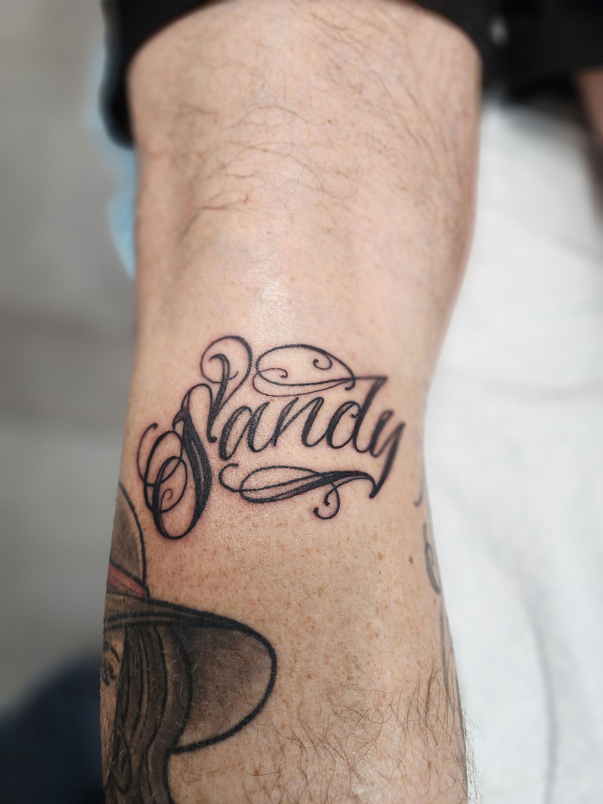 A man has a tattoo of the name sandy on his leg