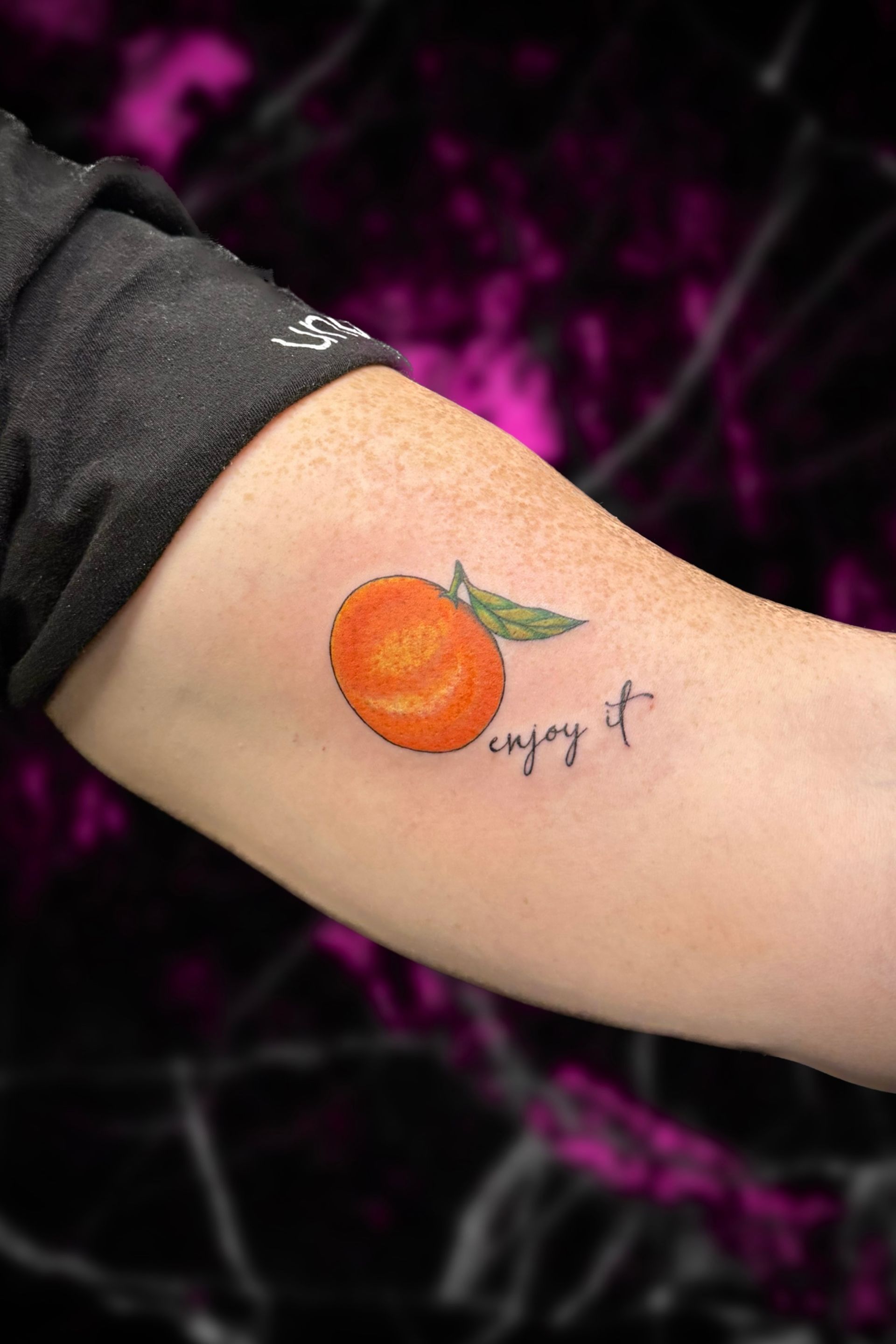 A person has a tattoo of an orange and the words enjoy it on their arm