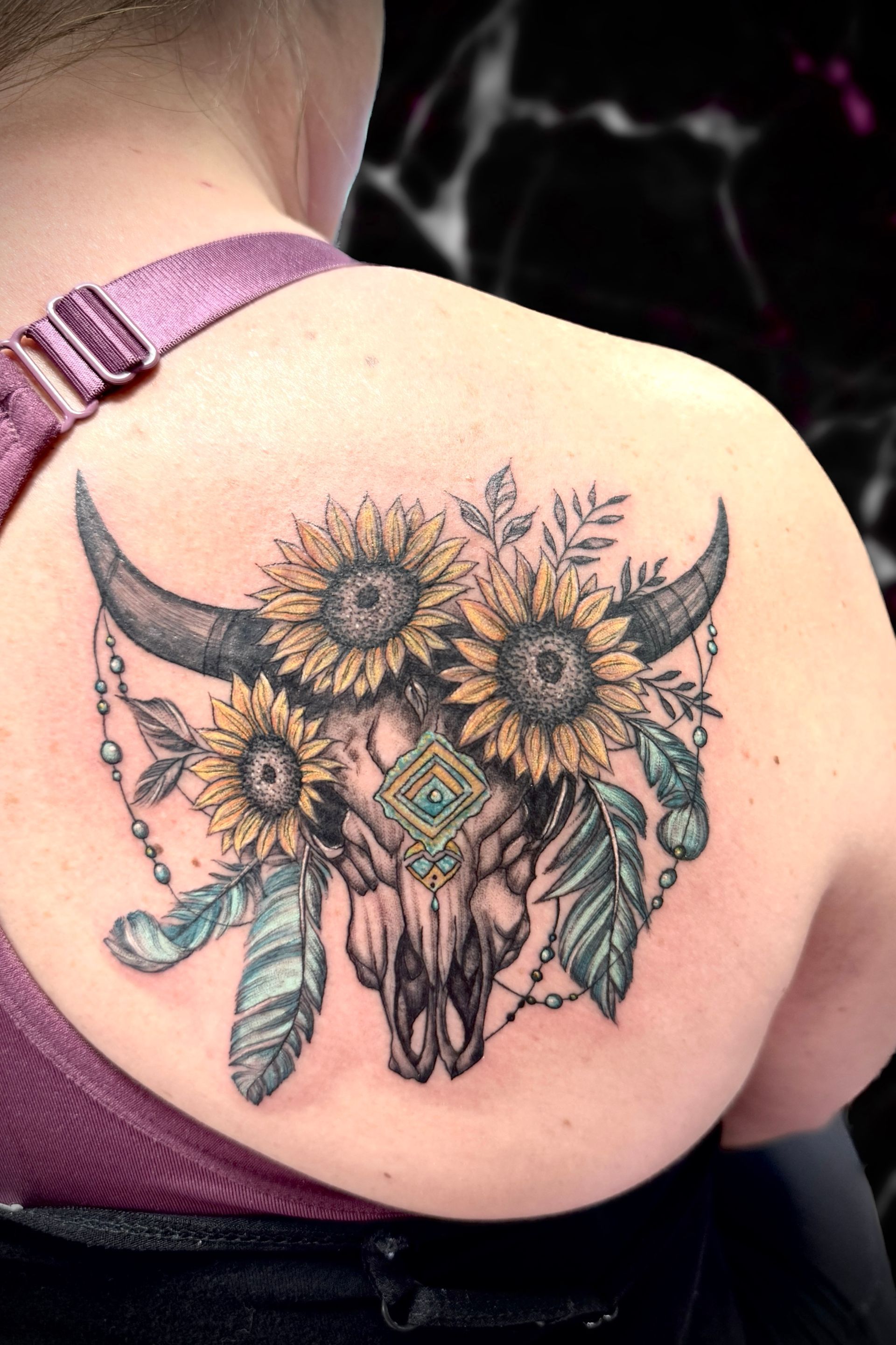 A woman has a tattoo of a bull skull with sunflowers and feathers on her shoulder
