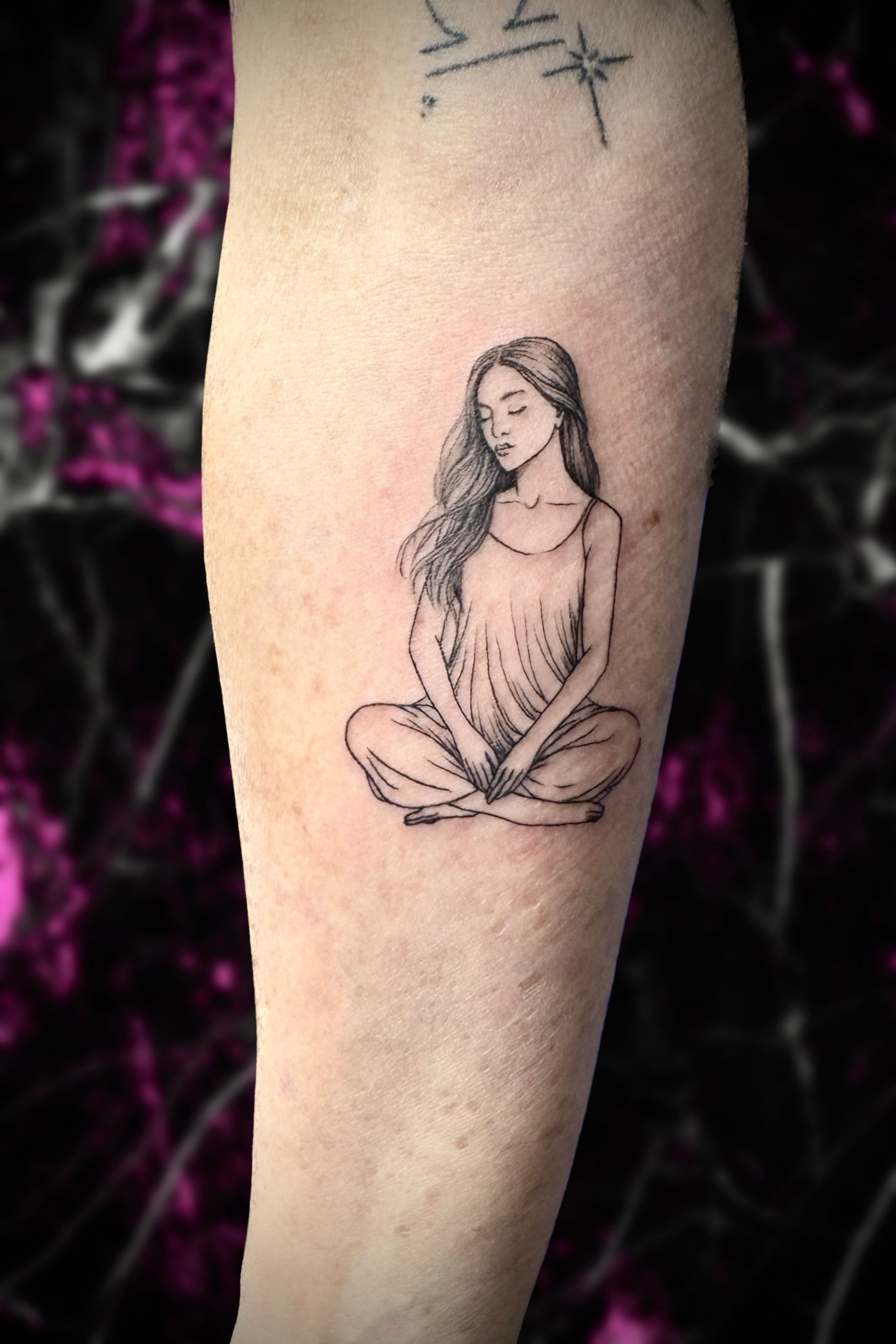 A woman has a tattoo of a woman sitting in a lotus position on her forearm