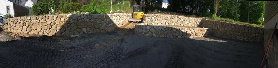 Freshly paved dark driveway with stone retaining walls and a paving machine in the distance