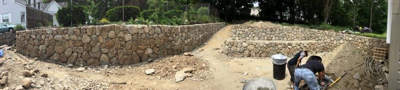 Workers laying stones in a large dirt excavation beside a stone retaining wall.