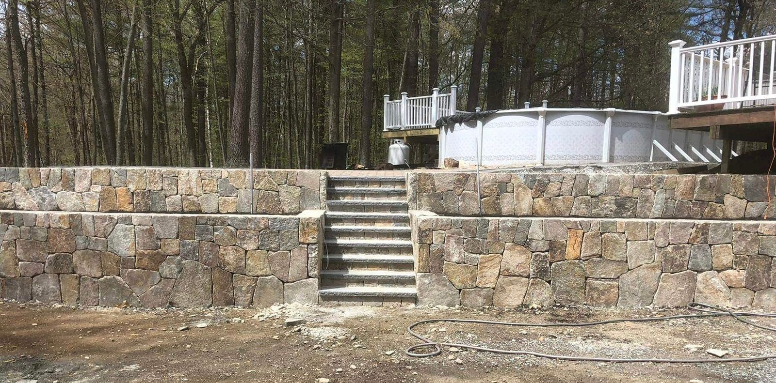 Stone retaining wall with central stairs leading to an above-ground pool in a wooded yard