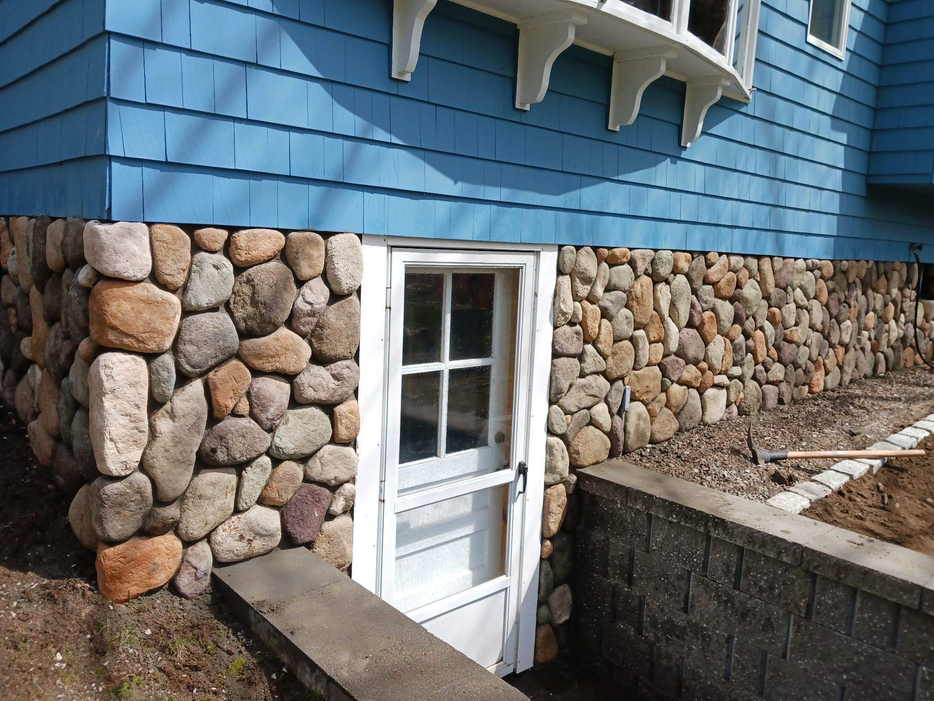 White door set in stone foundation beneath blue shingle siding and a small window.