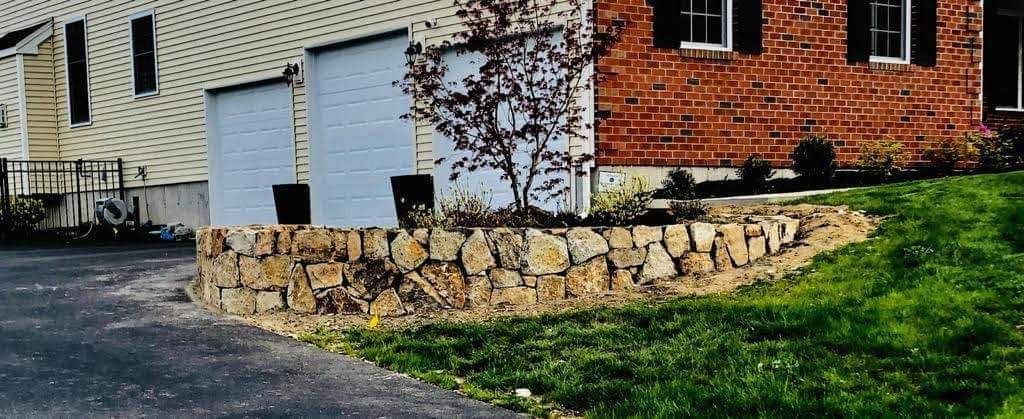 Stone retaining wall beside a driveway, with a white garage and red brick house in the background.