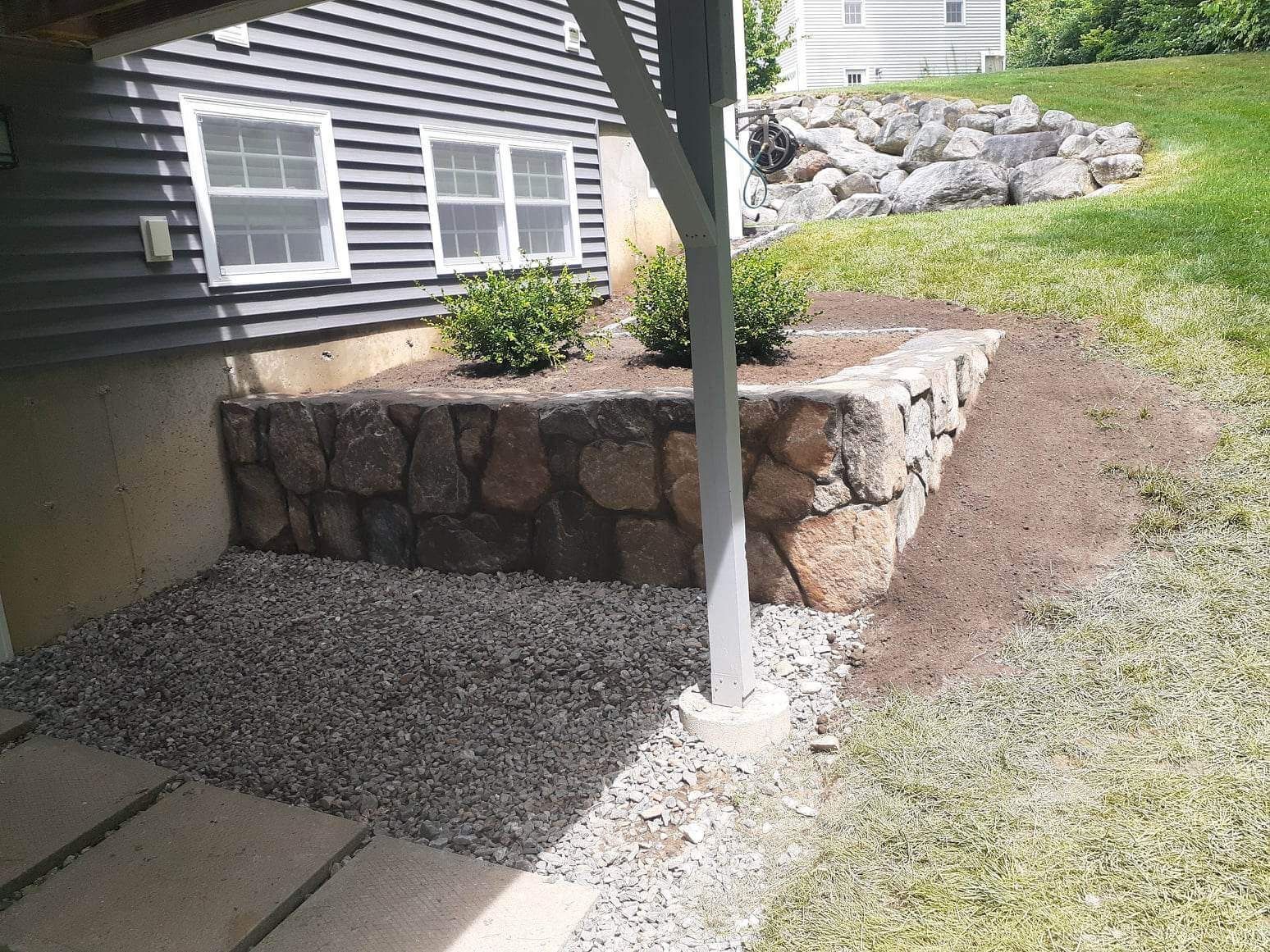 Gravel path beside a dark-sided house, with a stone retaining wall and white support post.