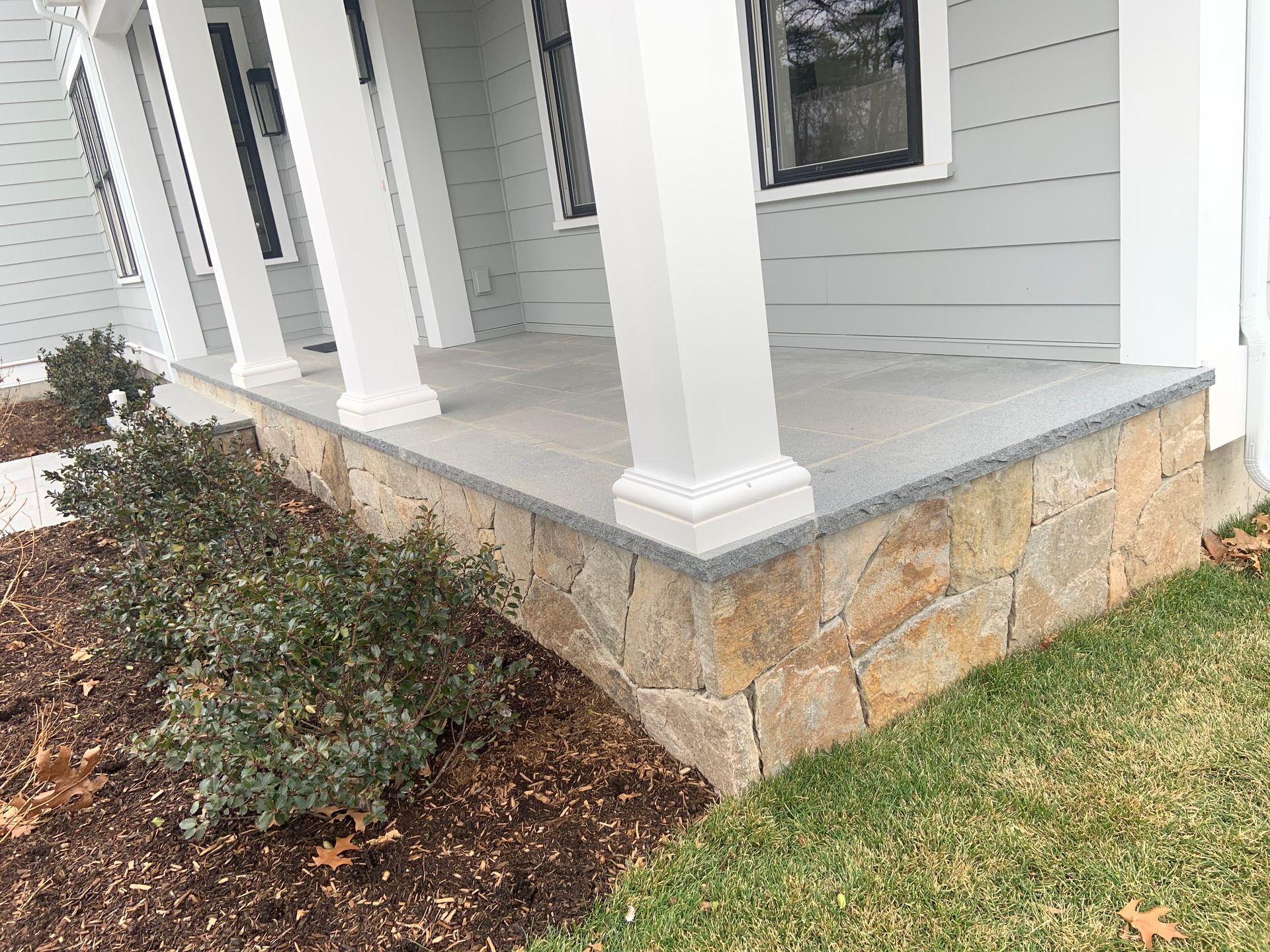 Front porch with white columns and stone base beside a shrub-lined lawn