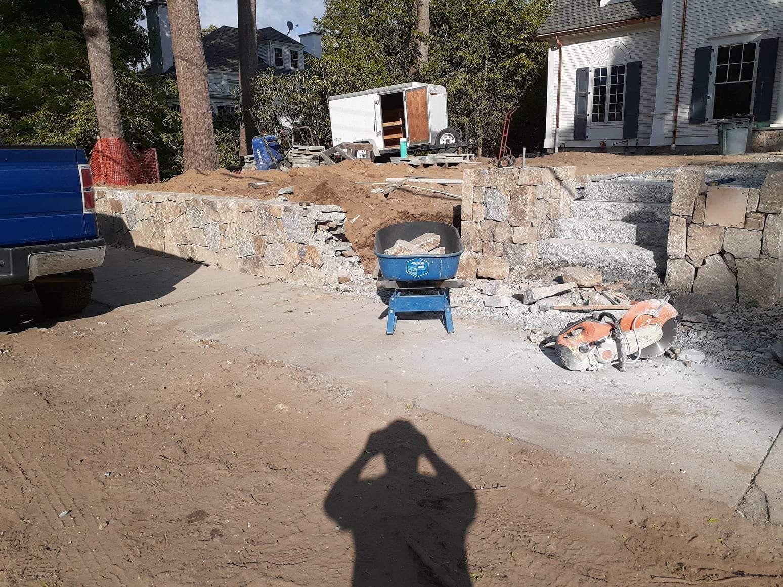 Construction site with a blue wheelbarrow, rubble, and a damaged stone wall beside a white building.