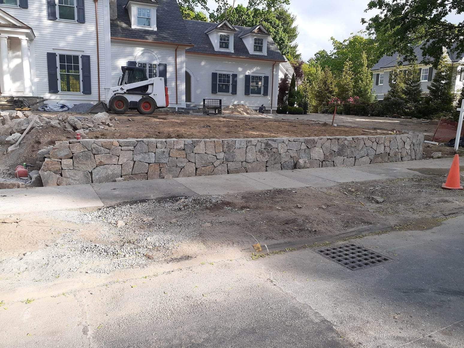 Stone retaining wall under construction in front of a house, with an orange cone and construction equipment.