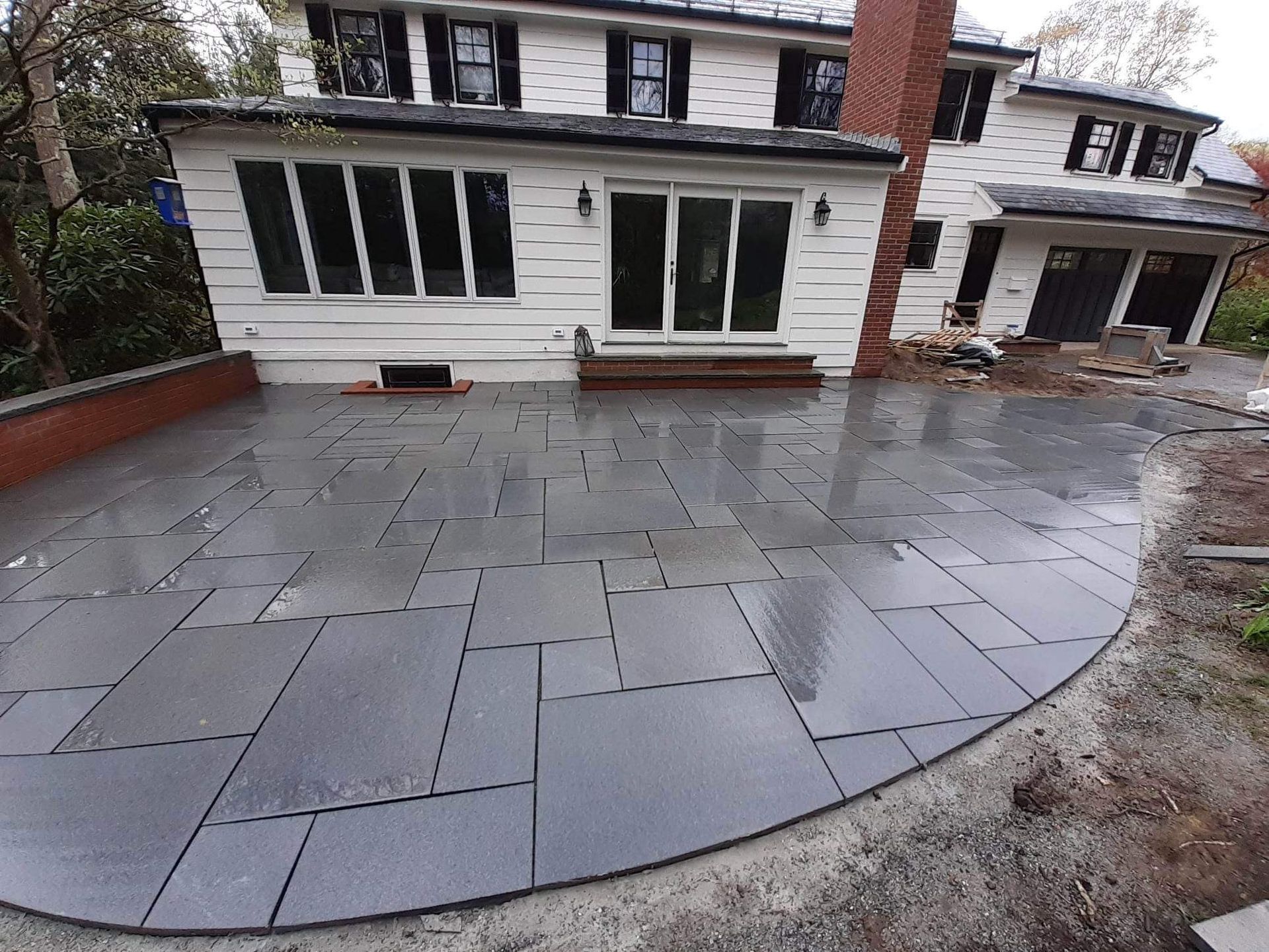 Curved gray stone patio in front of a white house with black-trimmed windows and a brick chimney.