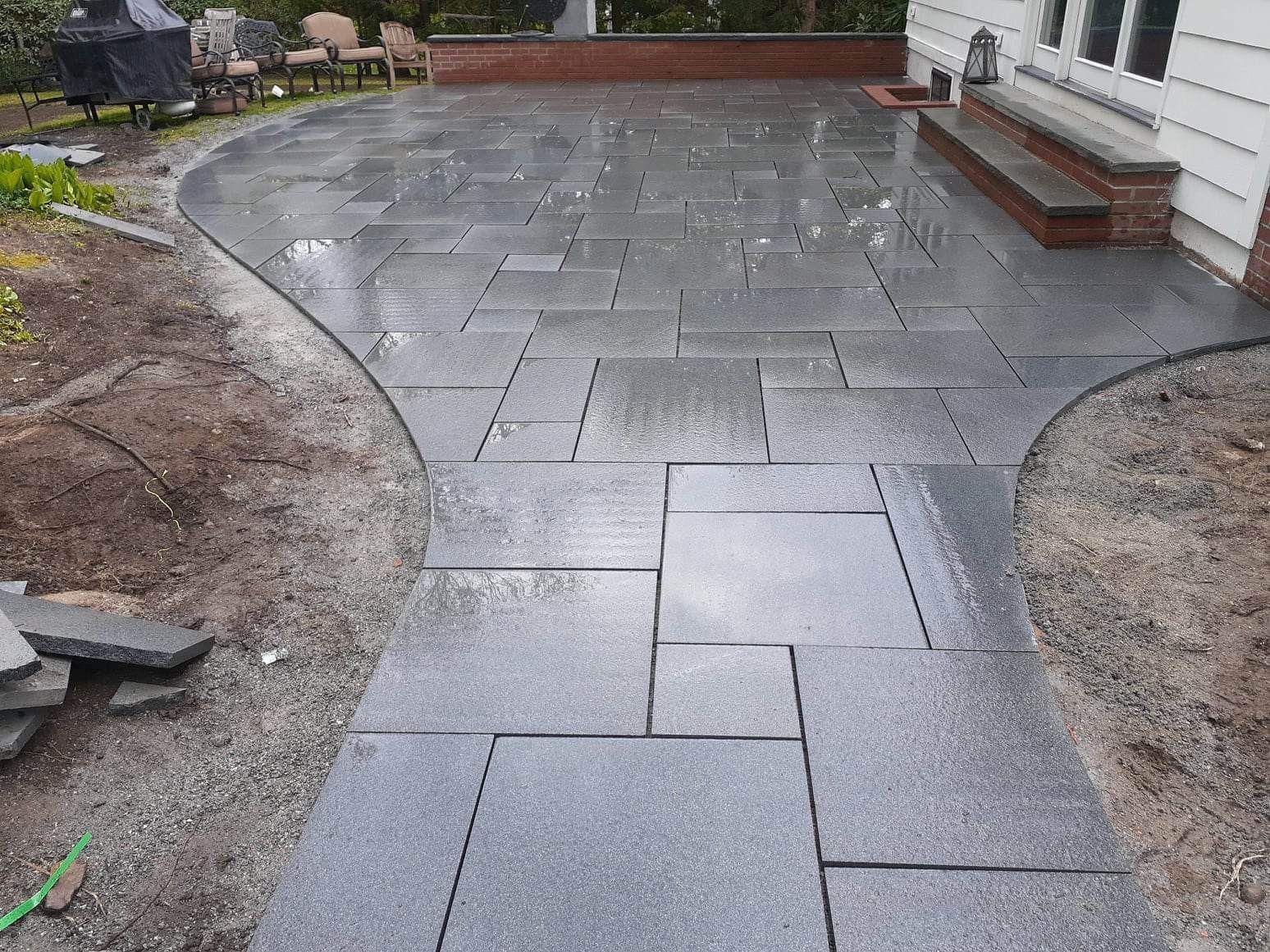 Curved gray stone patio under construction beside a white house, with soil edges and a brick retaining wall.