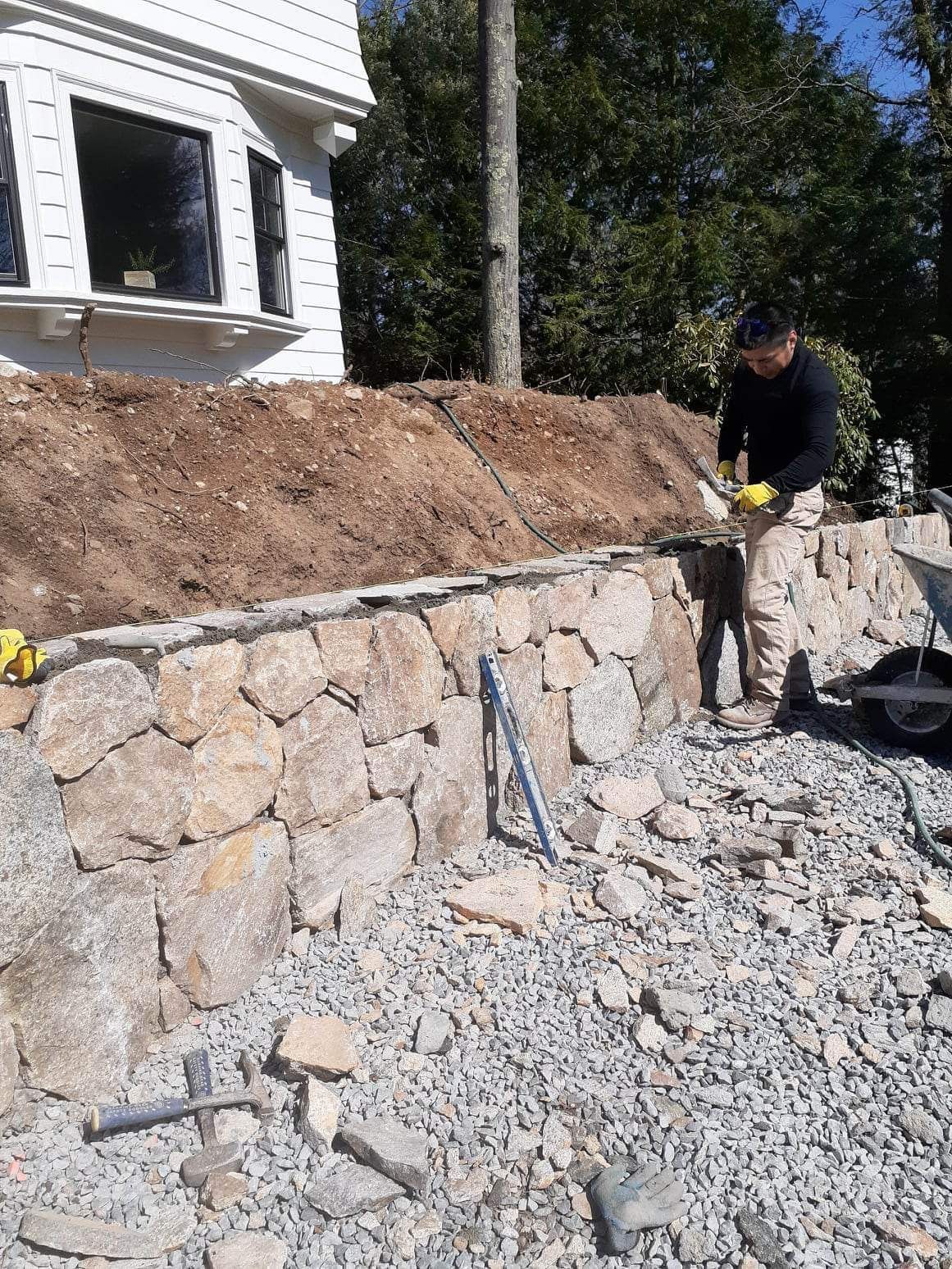 Worker building a stone retaining wall beside a house, with rubble and dirt piled around the site.