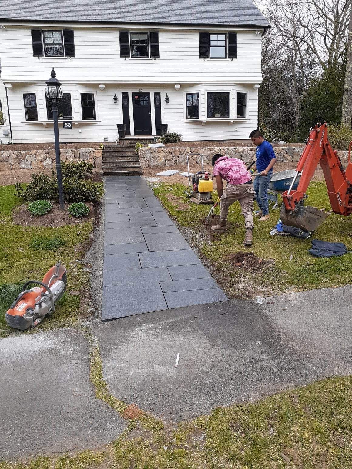 Workers laying a dark stone walkway in front of a white house with stone steps and a porch