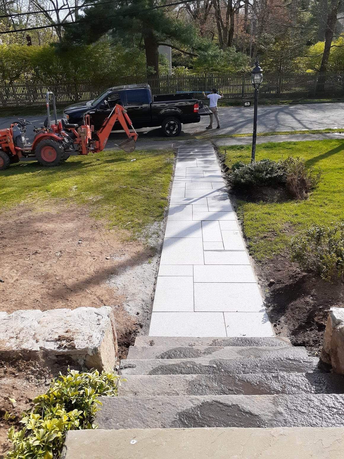Backyard walkway under construction with paving stones, a tractor, and a dump truck near a lawn and trees