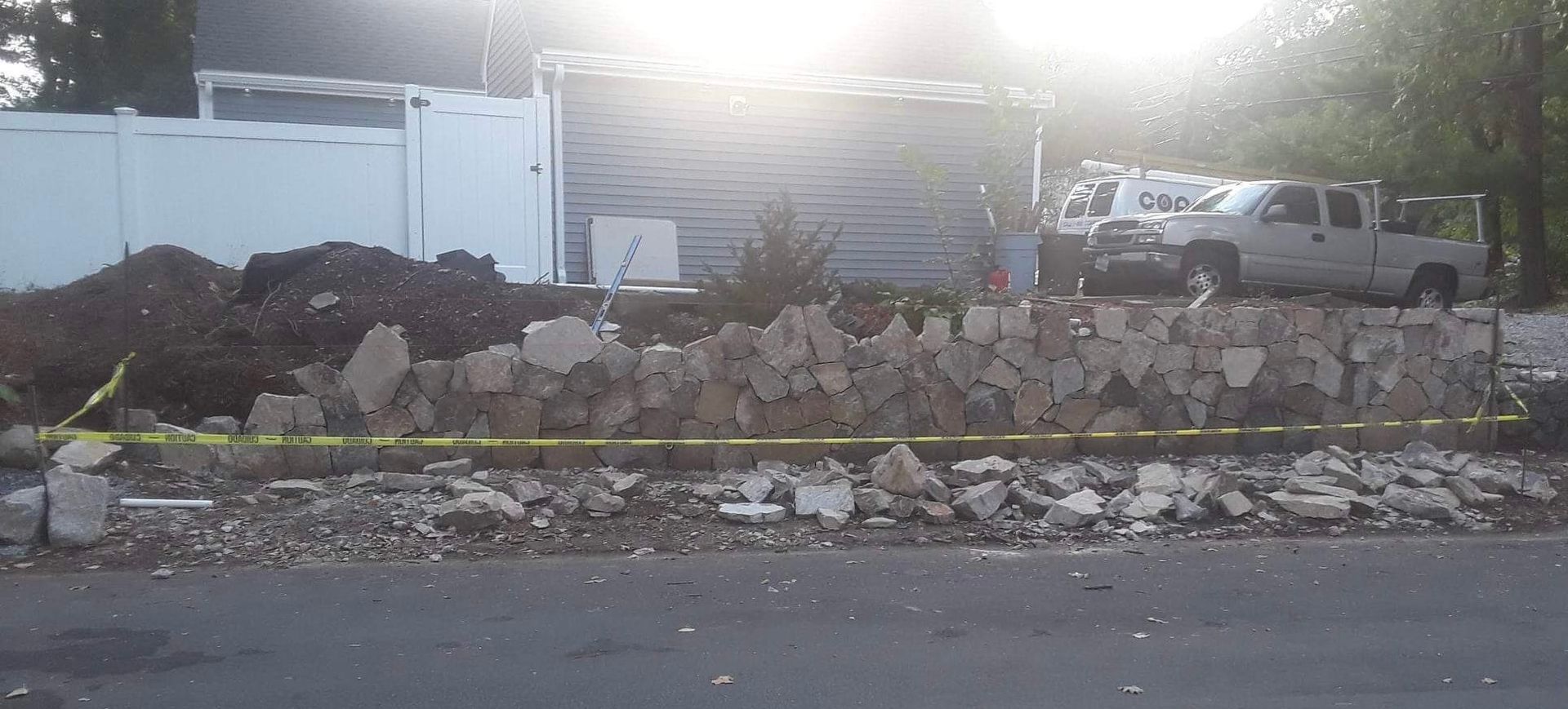 Sunlit roadside with a white pickup truck, rubble, and stacked materials behind a low stone wall