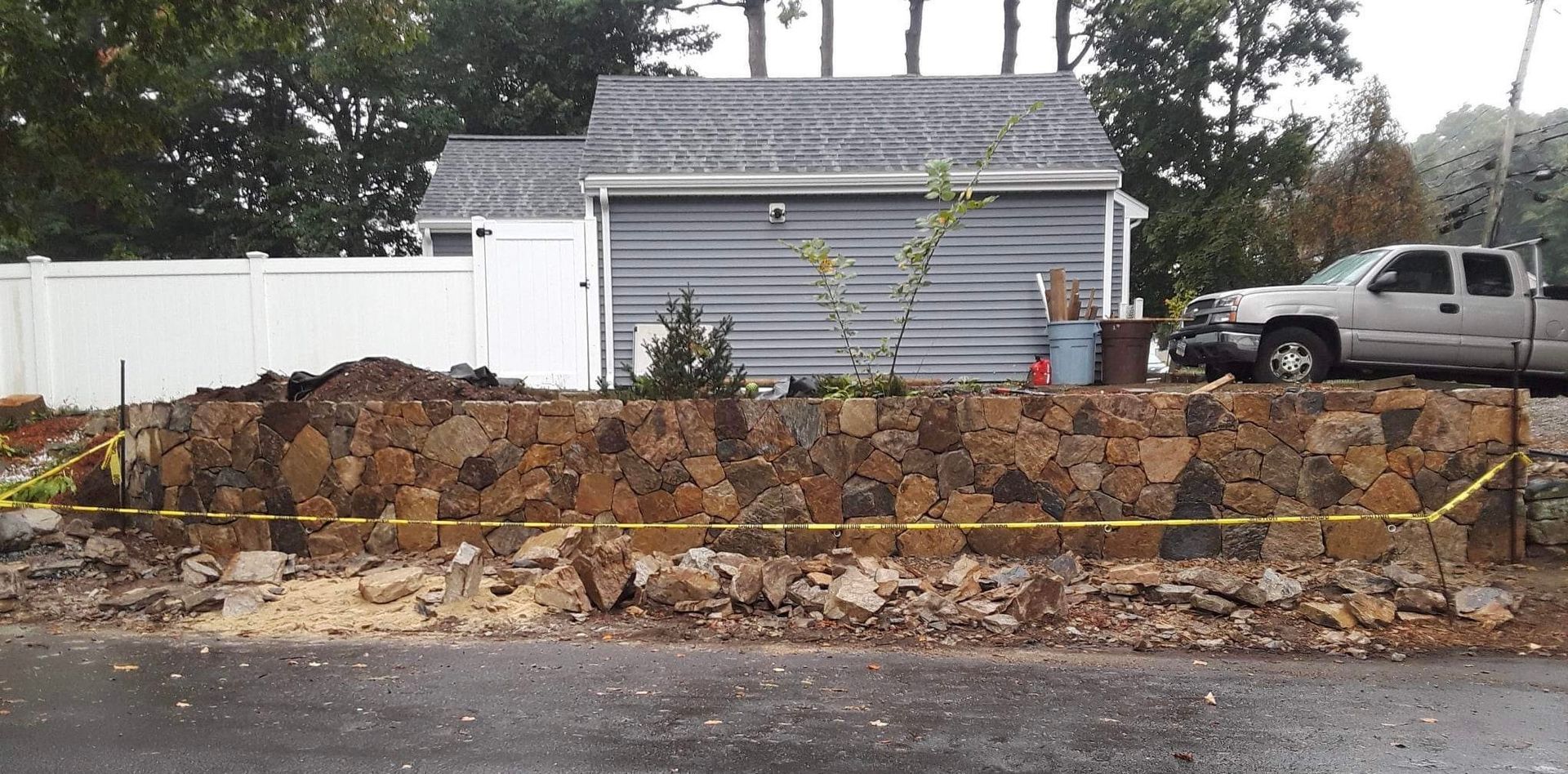 Excavation trench with dirt pile in front of a gray shed and parked pickup truck behind a white fence.