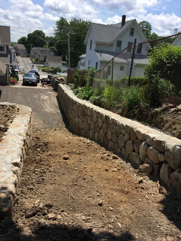 Stone retaining wall along a dirt construction path in a residential neighborhood with houses and a parked car.