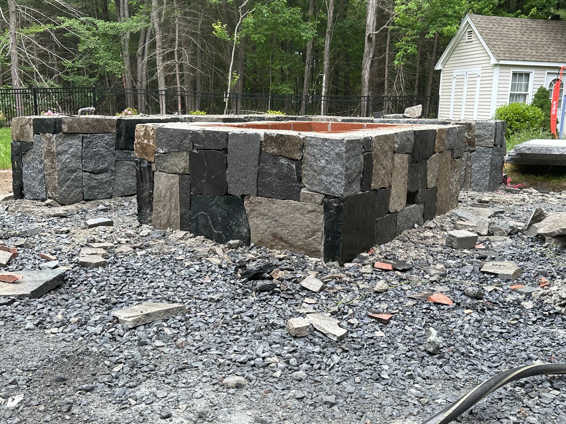 Stone fire pit under construction on a gravel patio beside a wooded yard and house