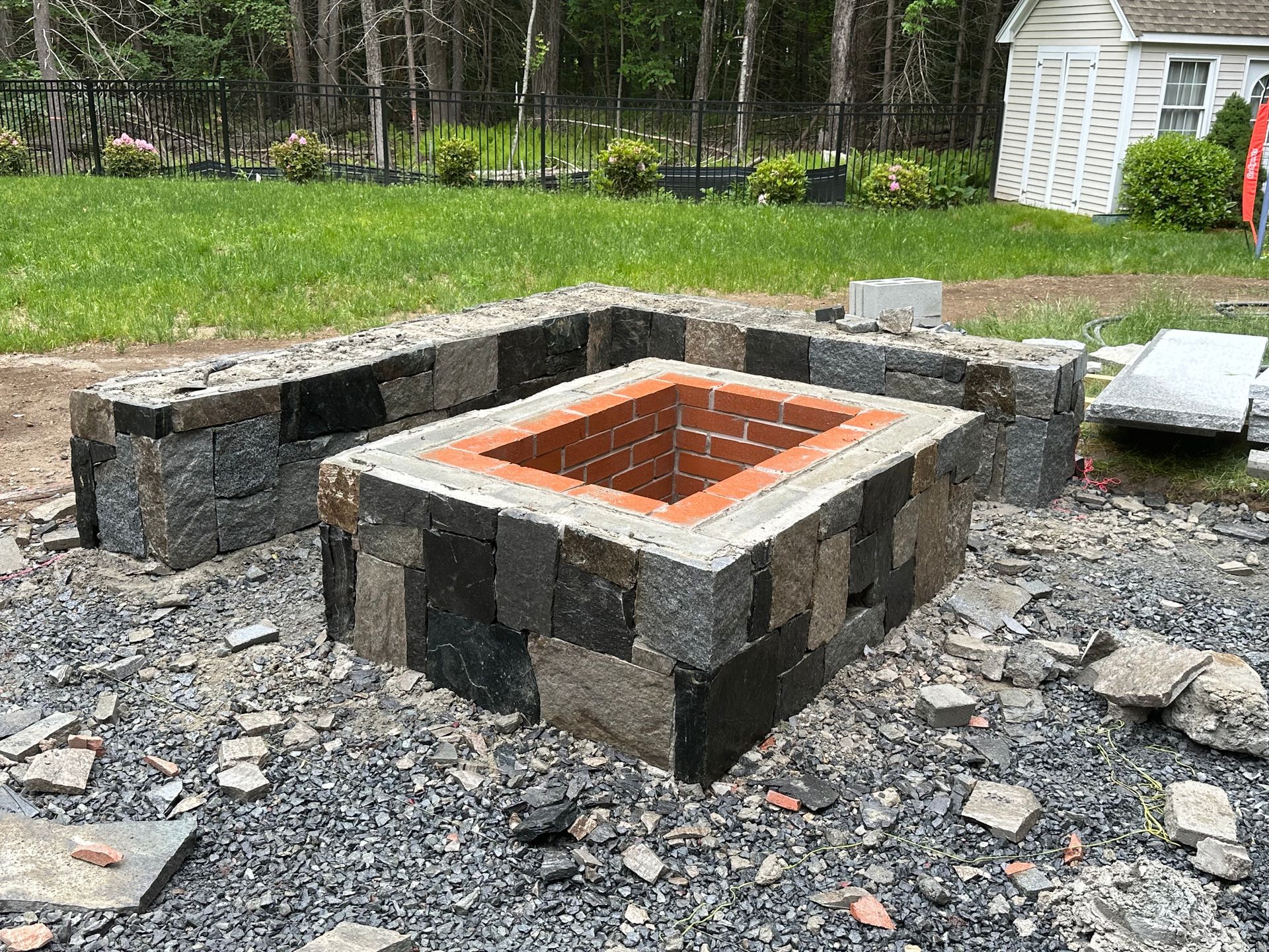 Stone fire pit under construction in a gravel yard, with a white shed in the background