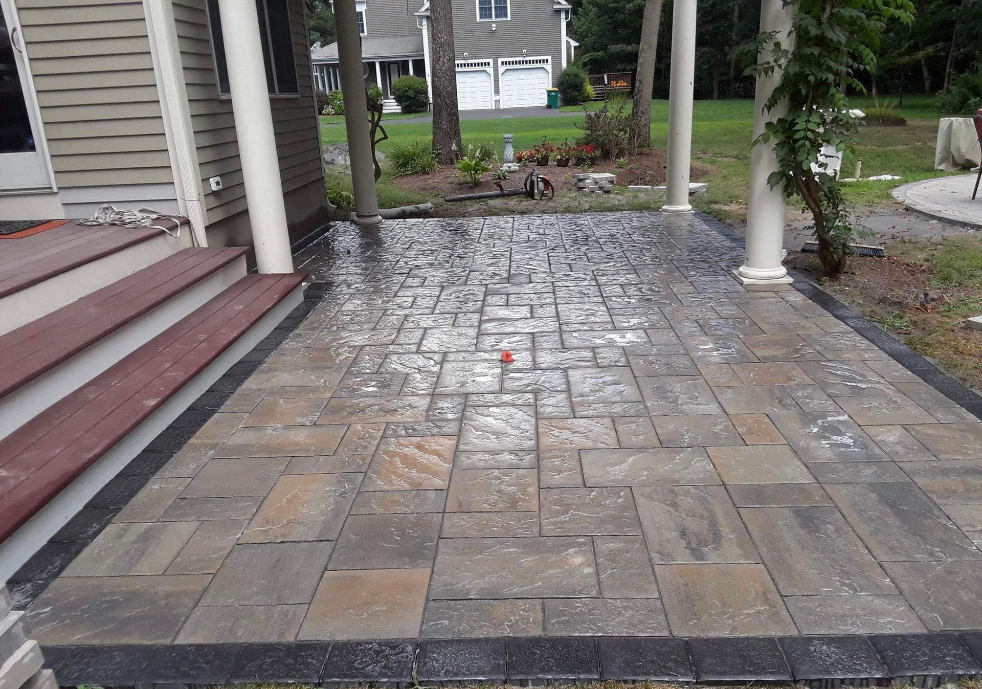 Wet stone patio beside house steps and white columns, with a garden path in the background.