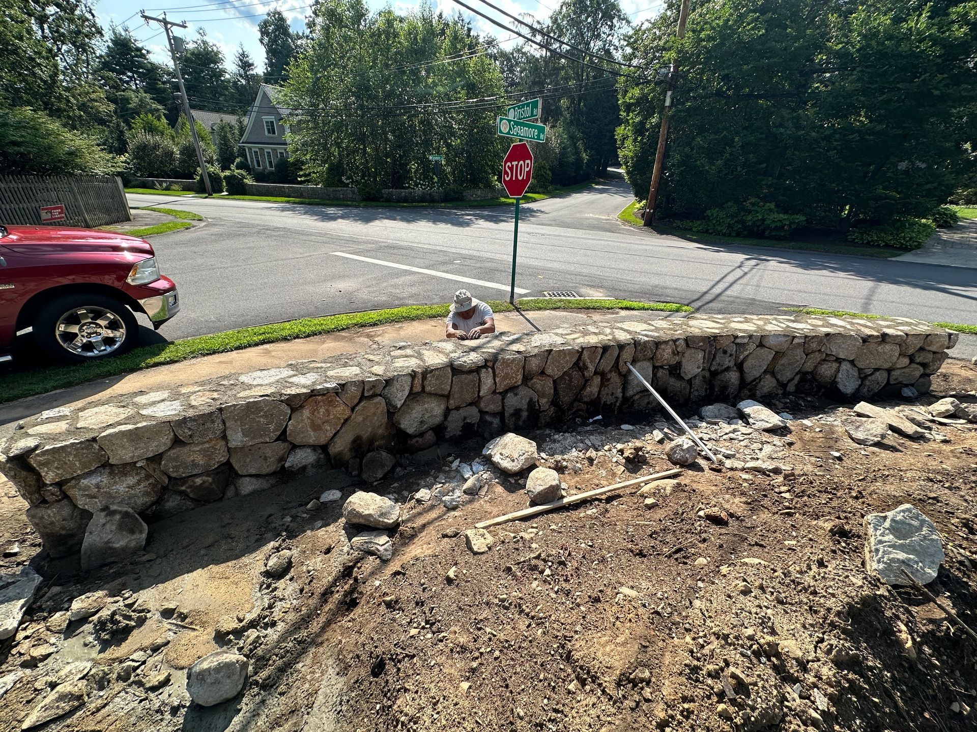 Stone retaining wall under construction beside a road, with a red stop sign and parked red car nearby.