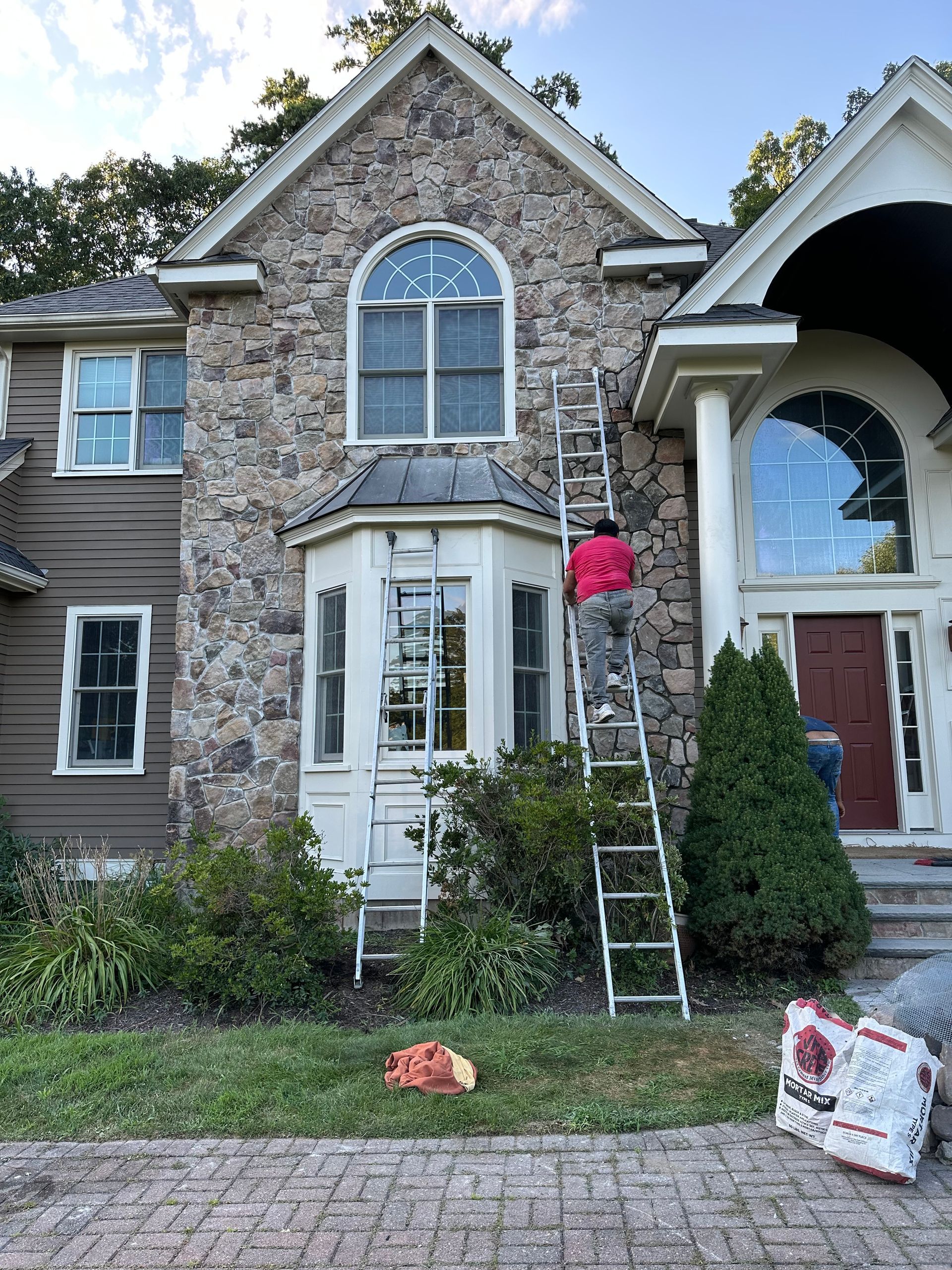 Stone townhouse front with white bay window, pink flowers, and a ladder leaning by the entrance.