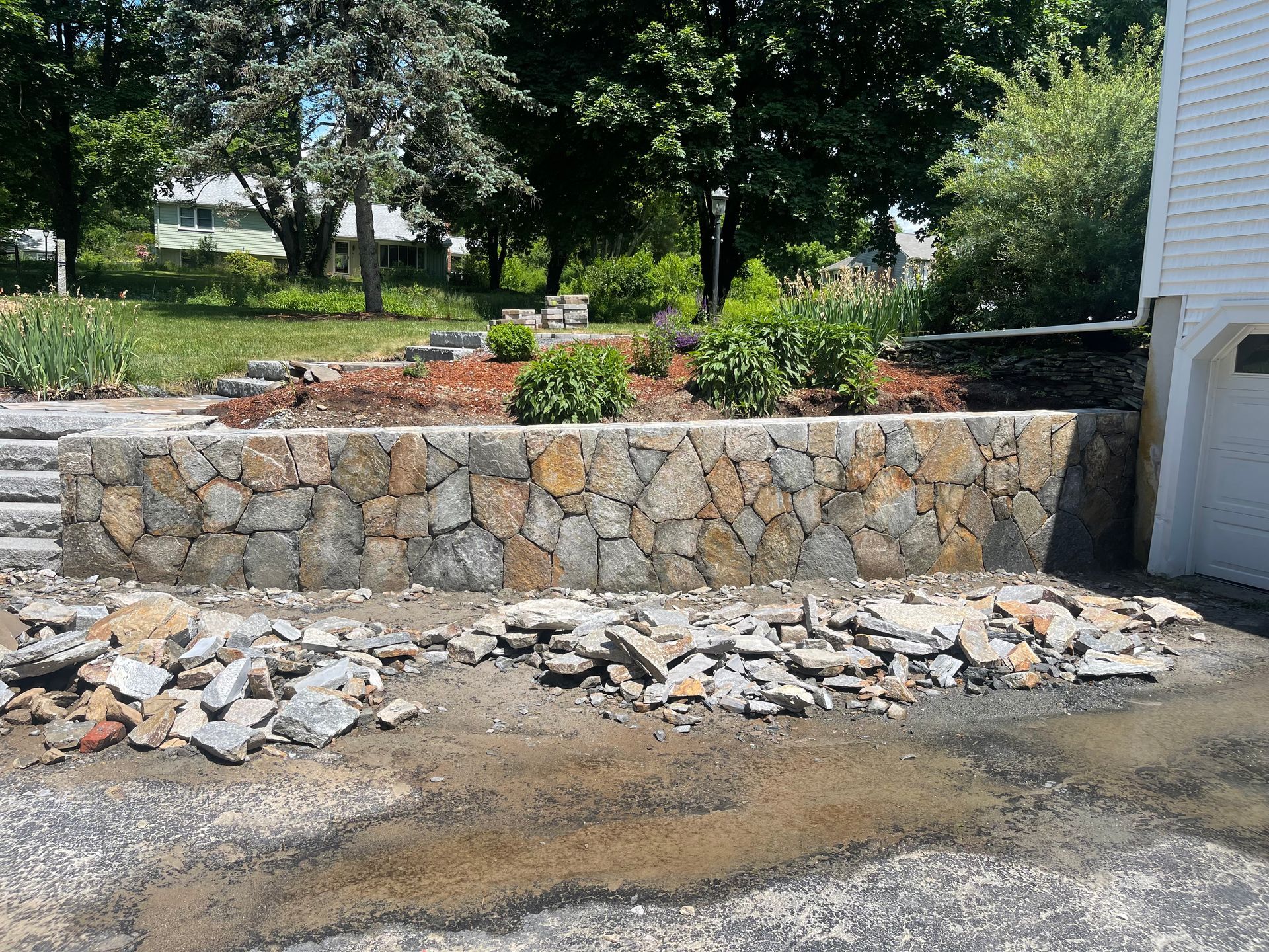 Stone retaining wall with rubble and gravel in front of a house driveway, surrounded by trees and landscaping