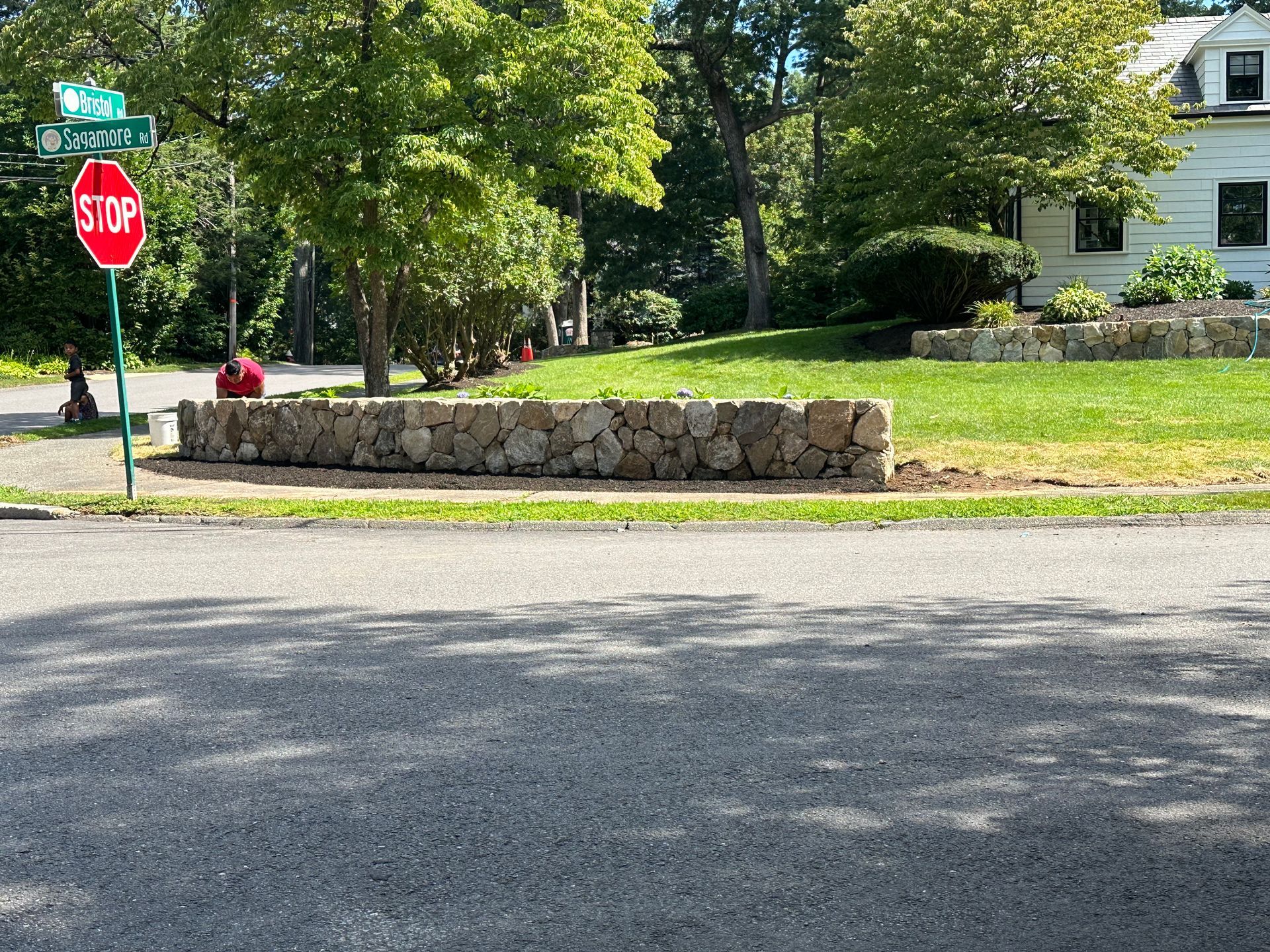 Quiet suburban street with a stop sign, stone wall, trees, and a white house in the background