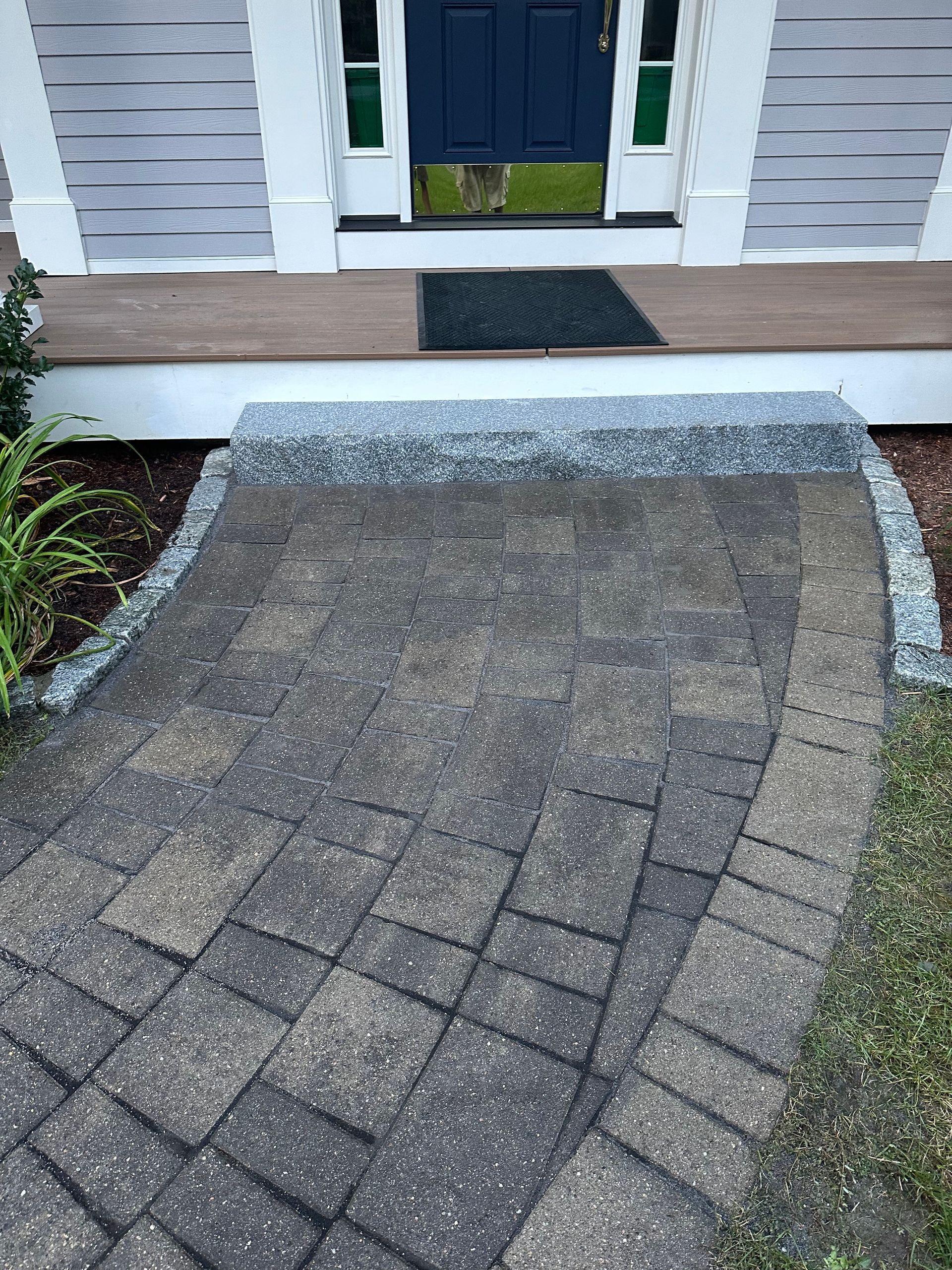 Brick walkway leading to a front door with a step and dark doormat, showing patchy surface wear