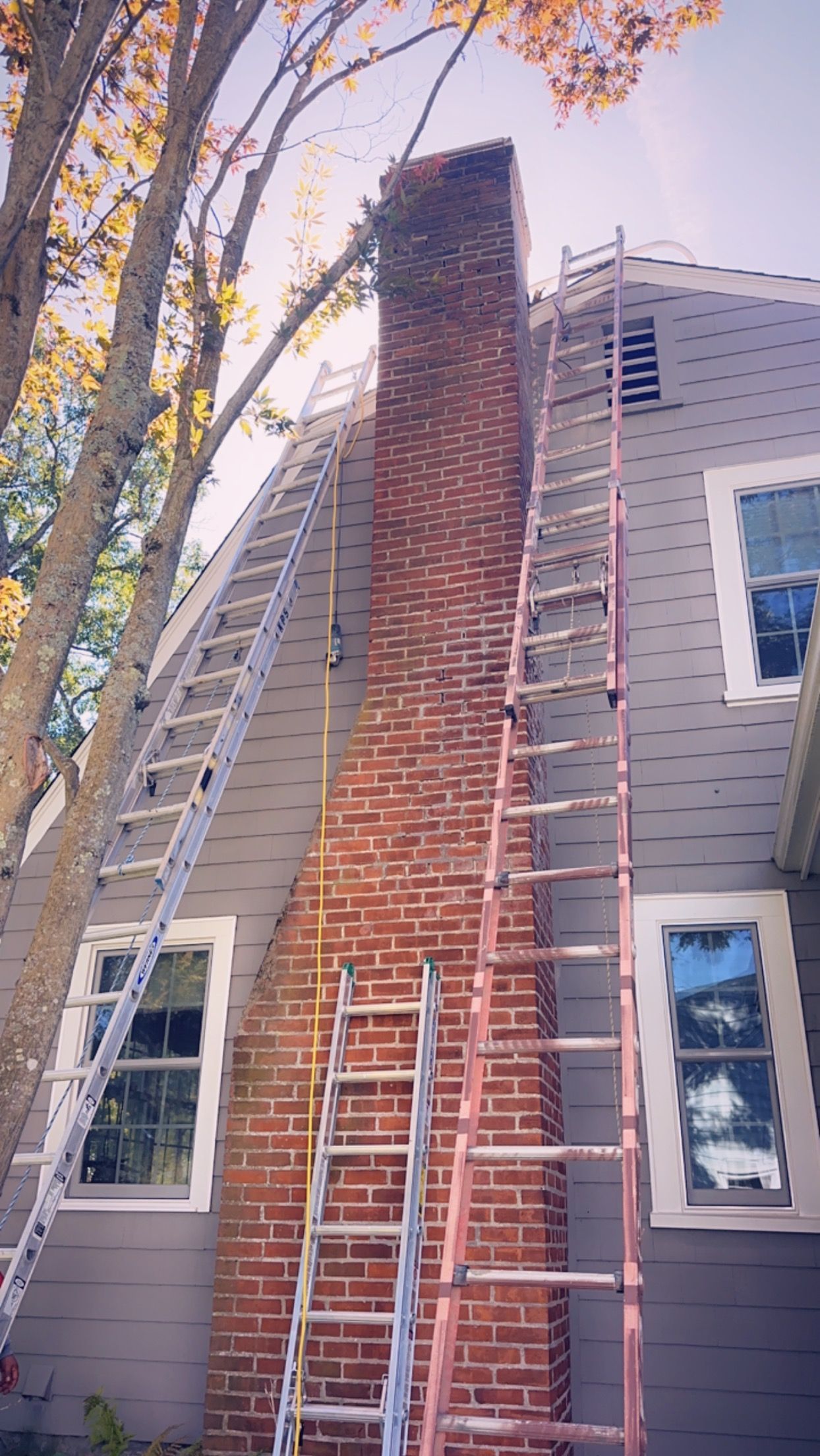 A tall brick chimney under construction beside a house, with two ladders and scaffolding against gray siding.