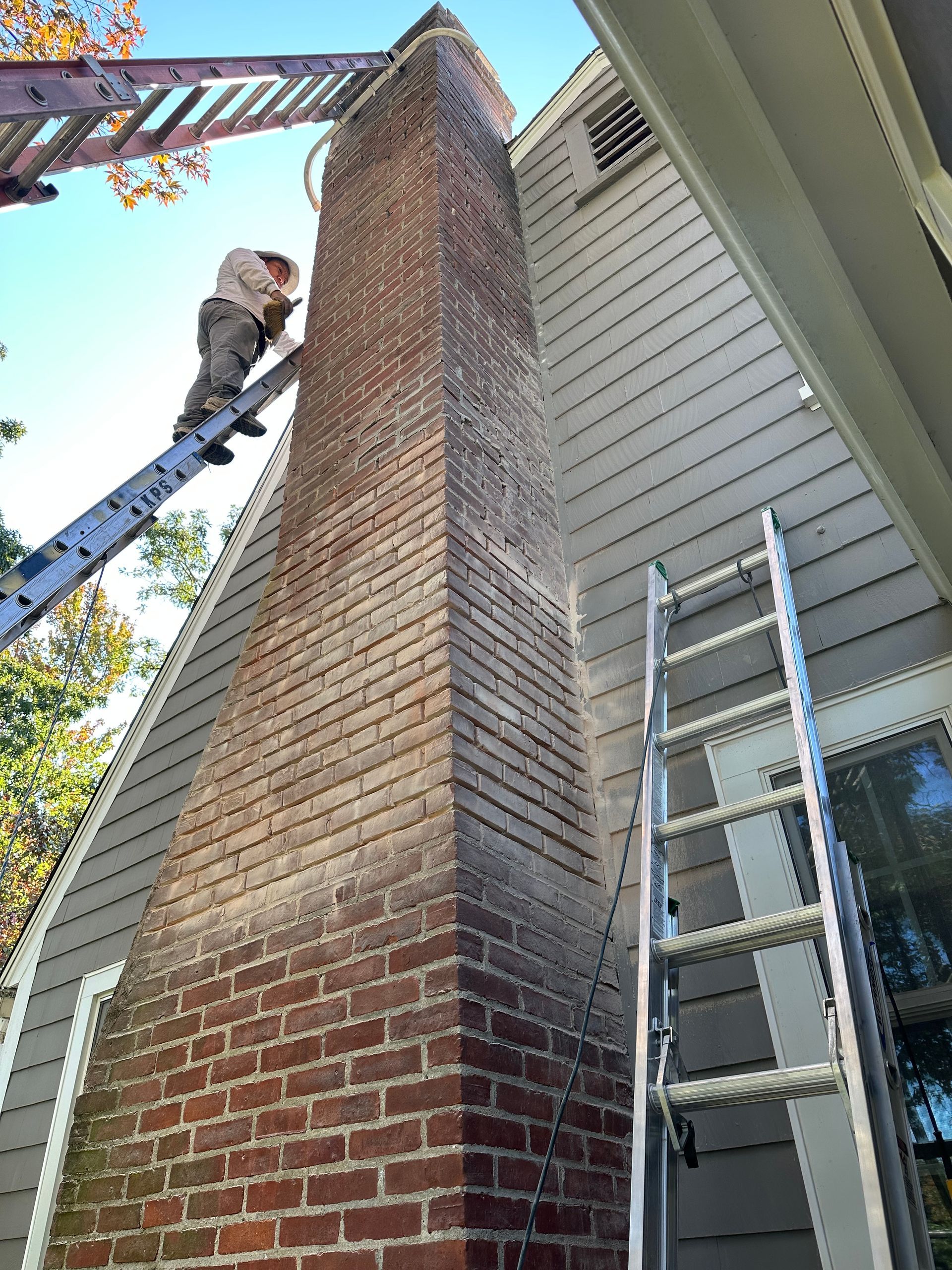 Worker on a ladder inspecting a tall brick chimney beside a house