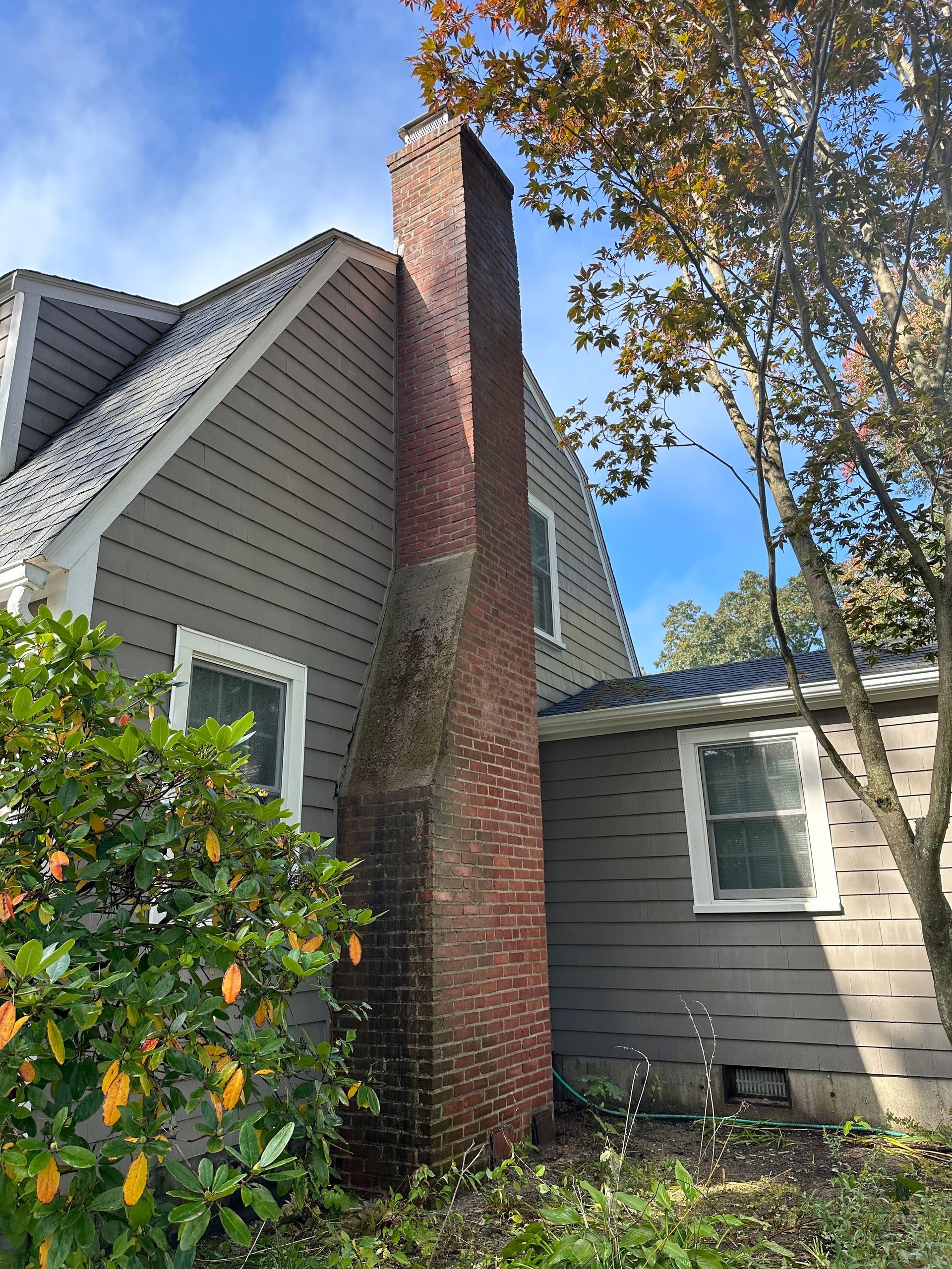 Brick chimney beside gray house with white-trimmed windows and a tree under a blue sky