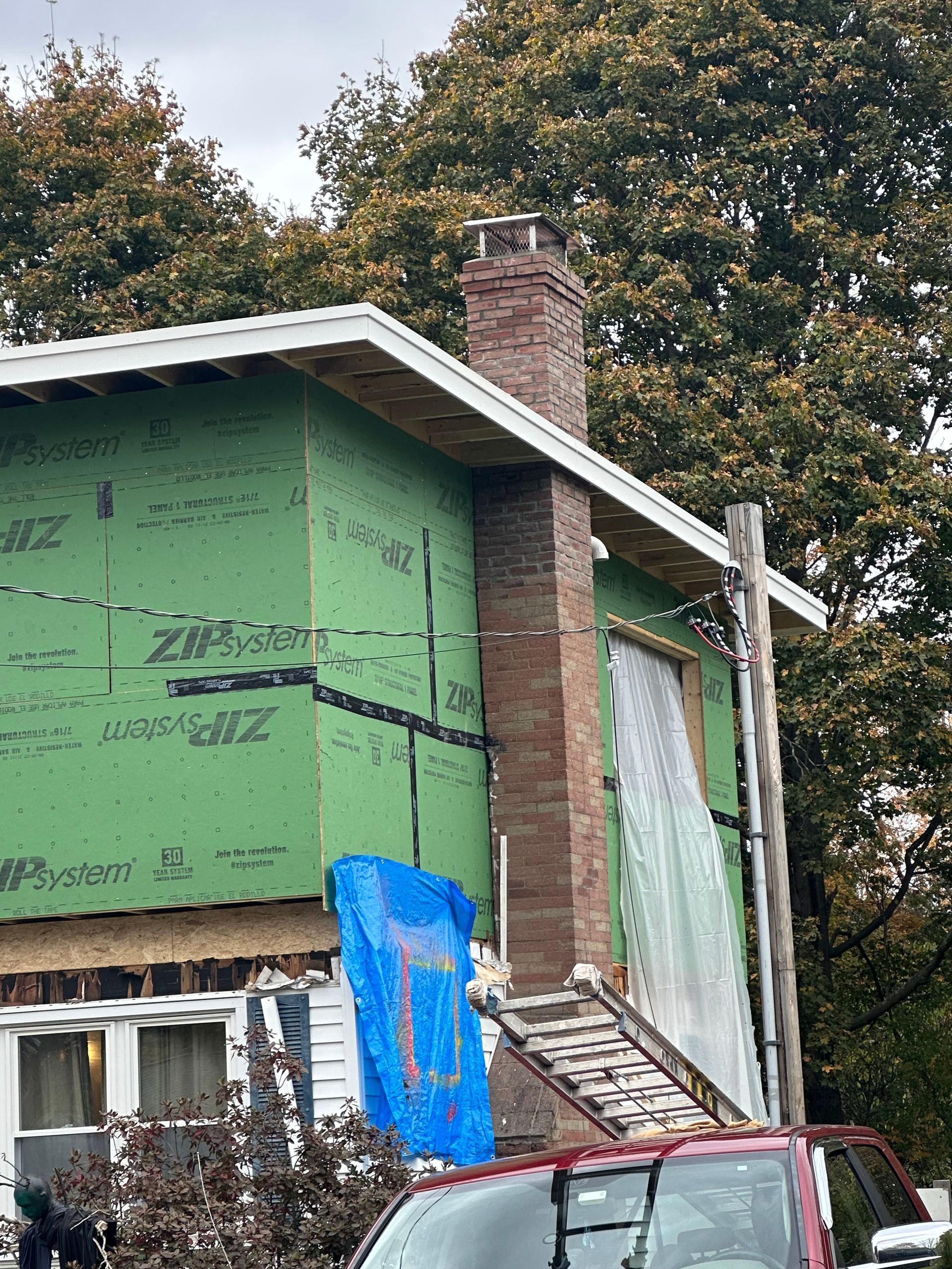 Partially renovated house with green insulation, brick chimney, and covered windows under trees