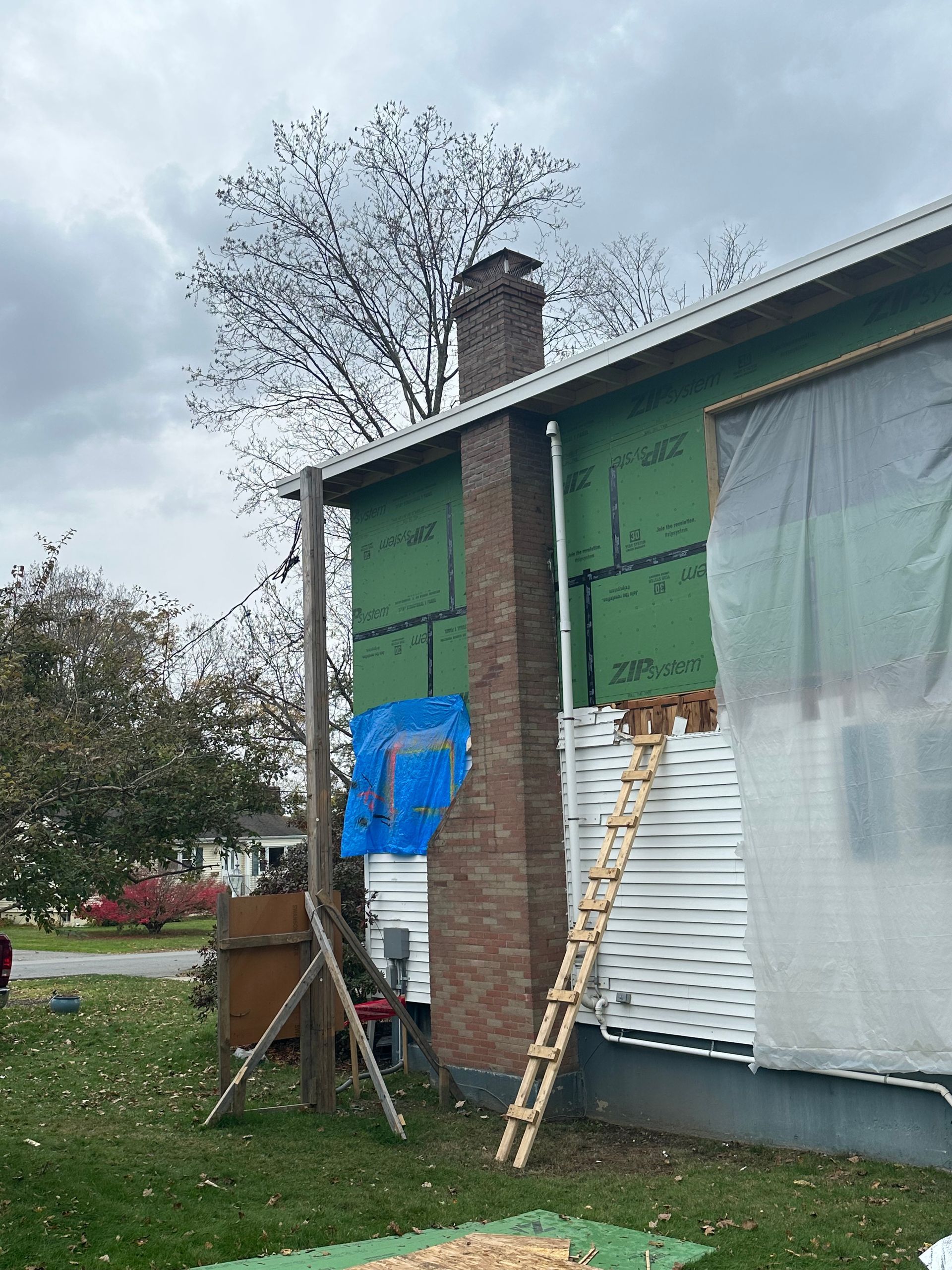House exterior under renovation with ladders, brick chimney, blue tarp, and covered windows