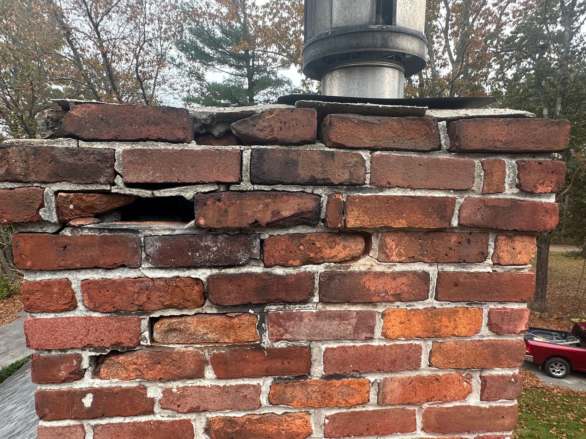 Damaged brick chimney with missing mortar and a small opening, outdoors with trees and a red car in the background