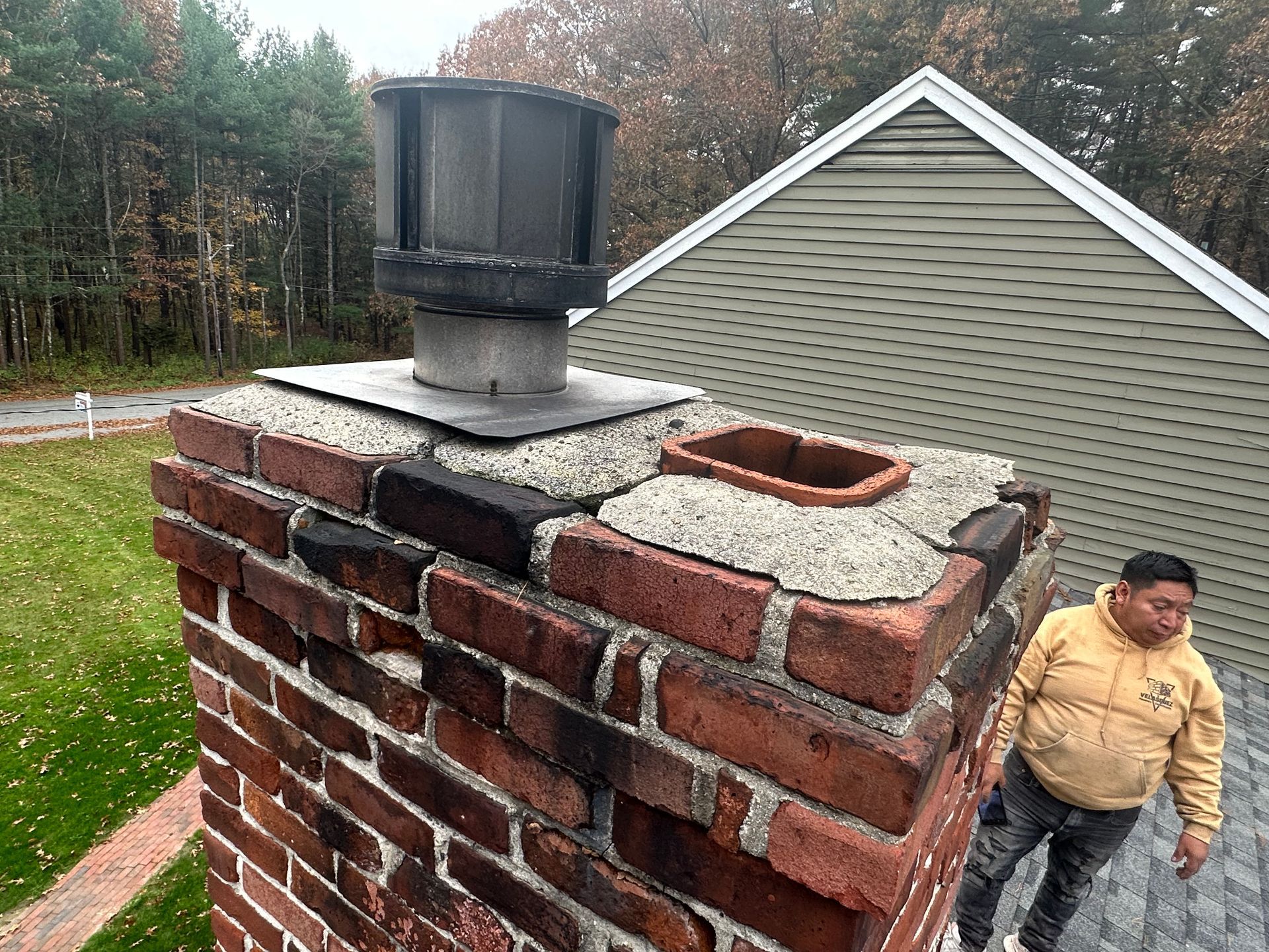 Brick chimney with a black vent cap beside a gray house, with a man standing nearby outdoors.