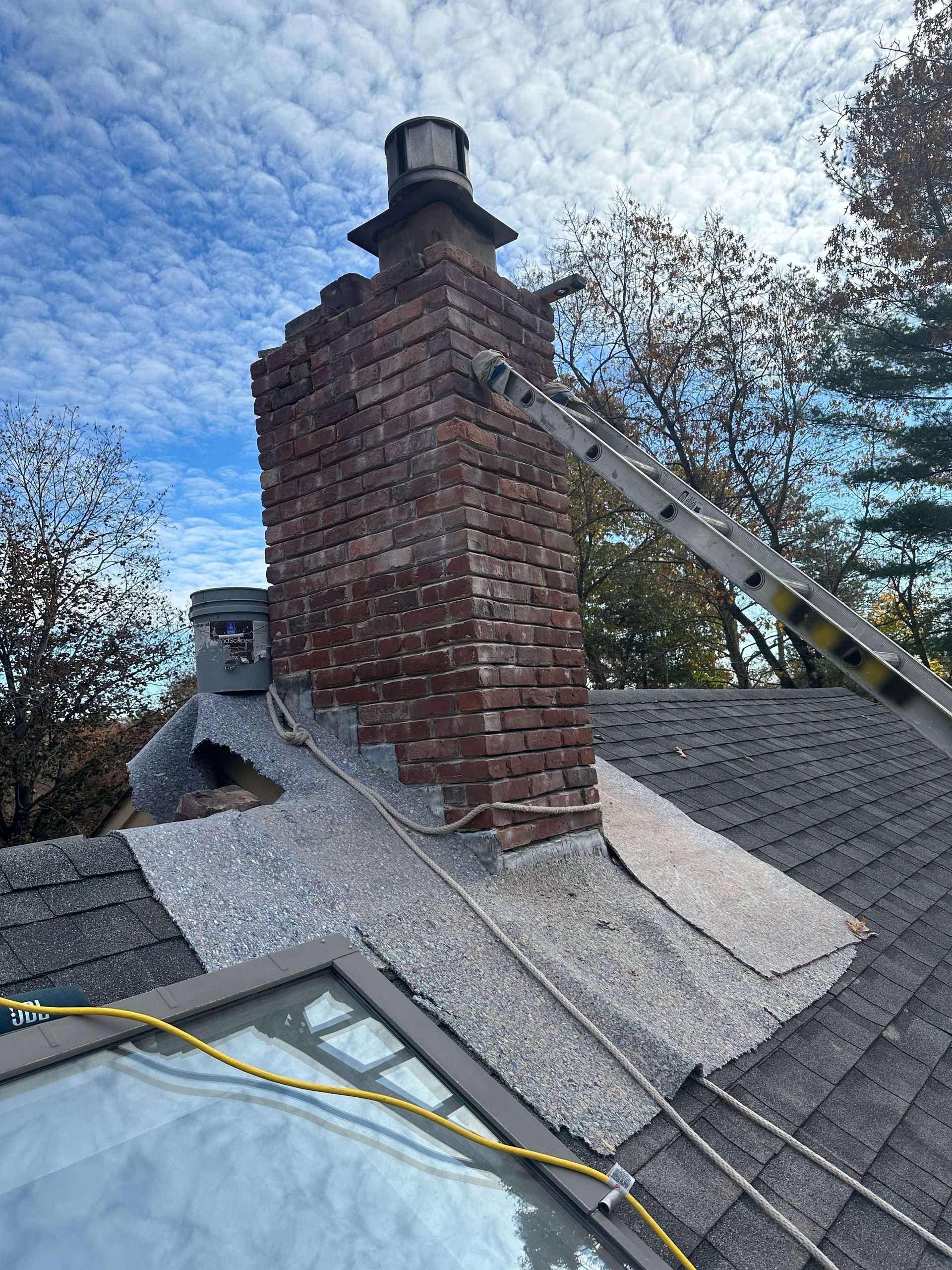 Brick chimney on a shingled roof, with a ladder leaning against it under a cloudy sky.