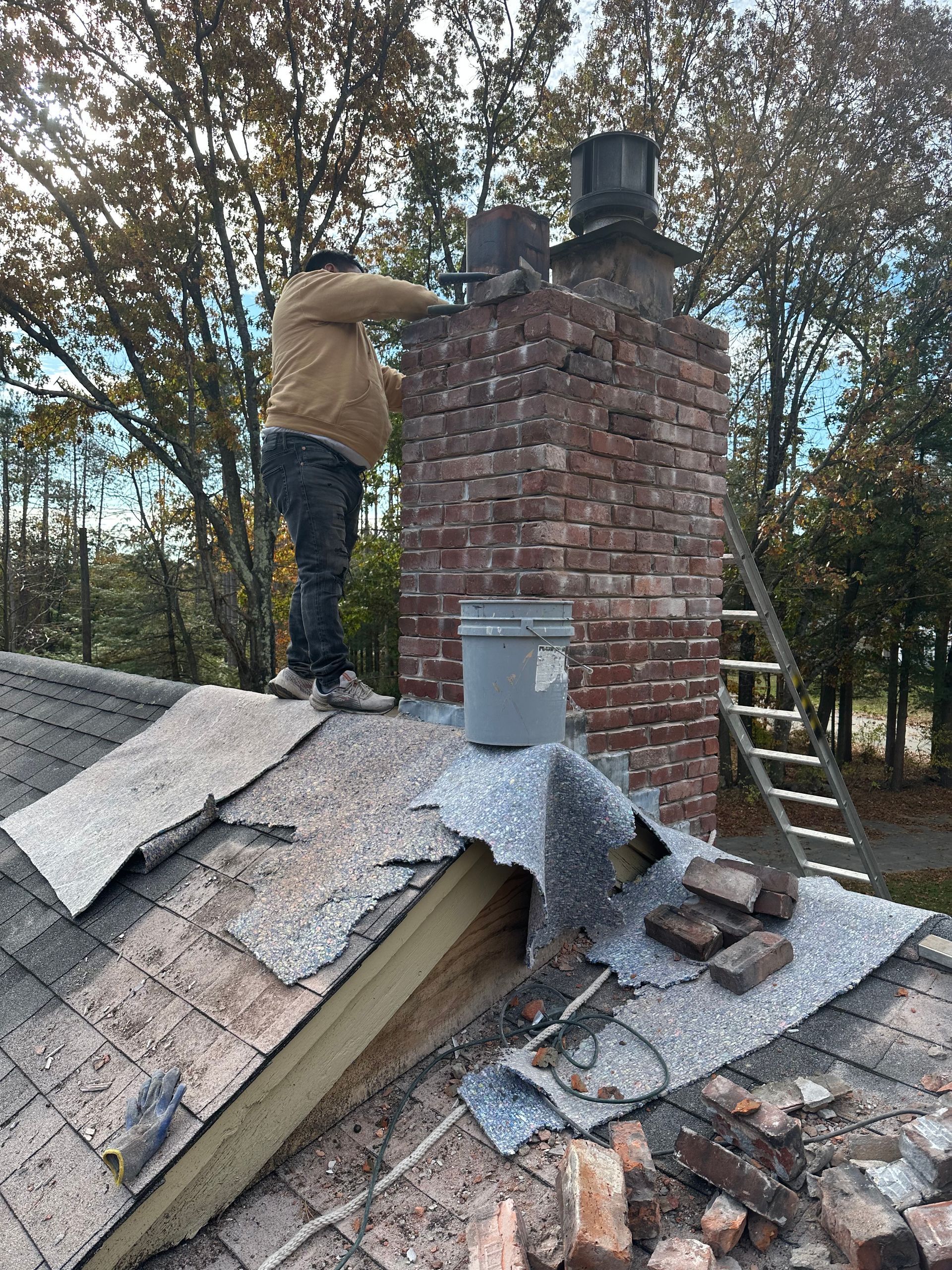 Person repairing a brick chimney on a damaged roof with exposed shingles and debris