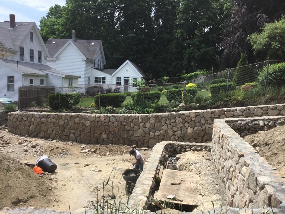 Stone retaining walls and a trench in a backyard under construction, with houses in the background