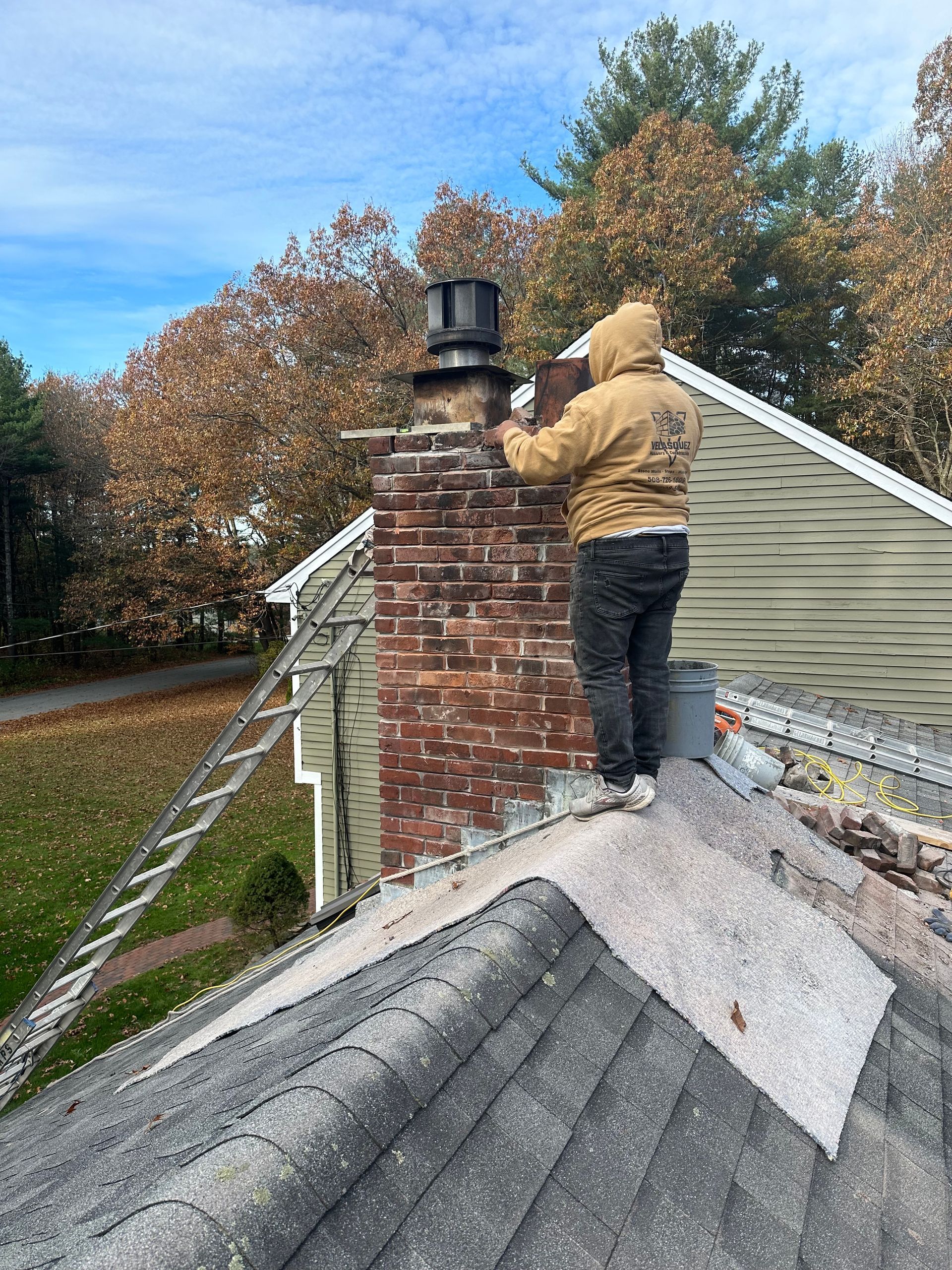Person painting a brick chimney on a shingled roof beside white siding and autumn trees