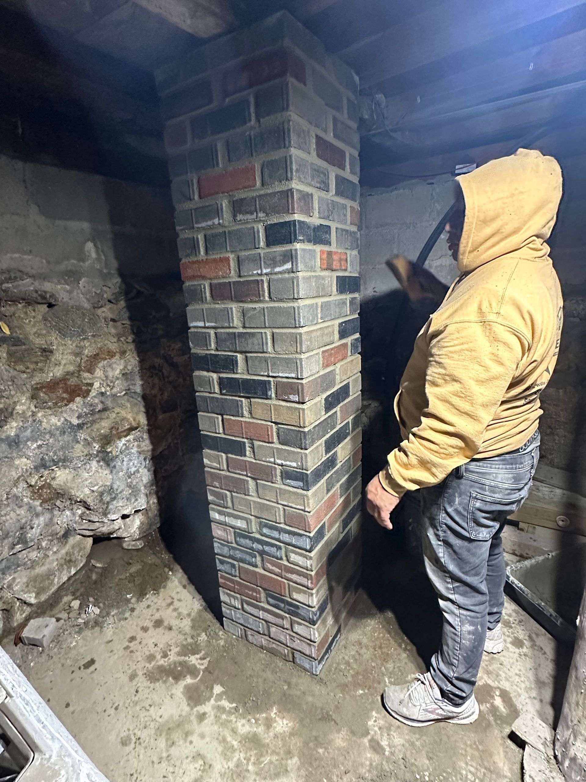 Person in a hooded jacket stands in a damp basement beside a brick chimney support pillar.