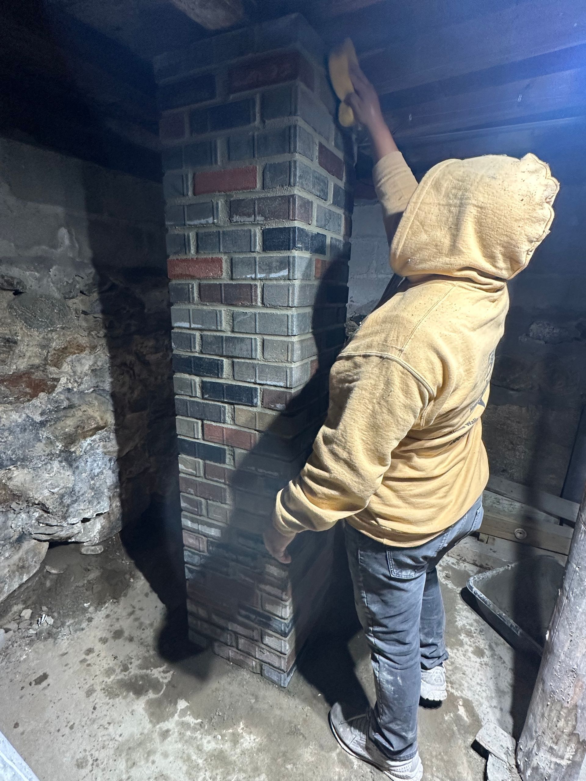 Person in a tan hoodie reaching up to inspect a brick chimney in a basement.