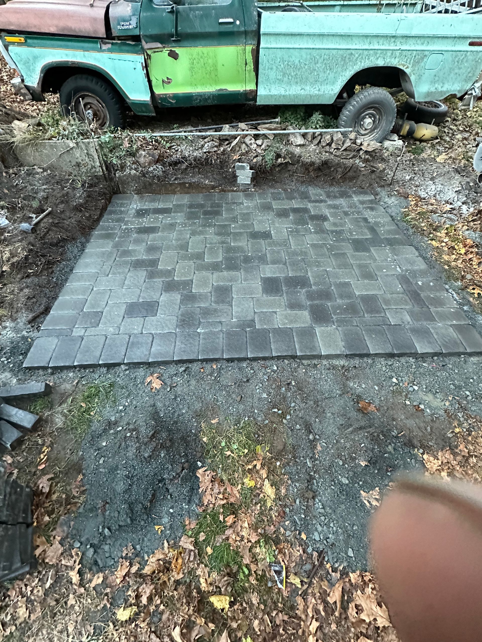 Fresh gray paver patio beside an old green pickup truck, with leaves and dirt around it