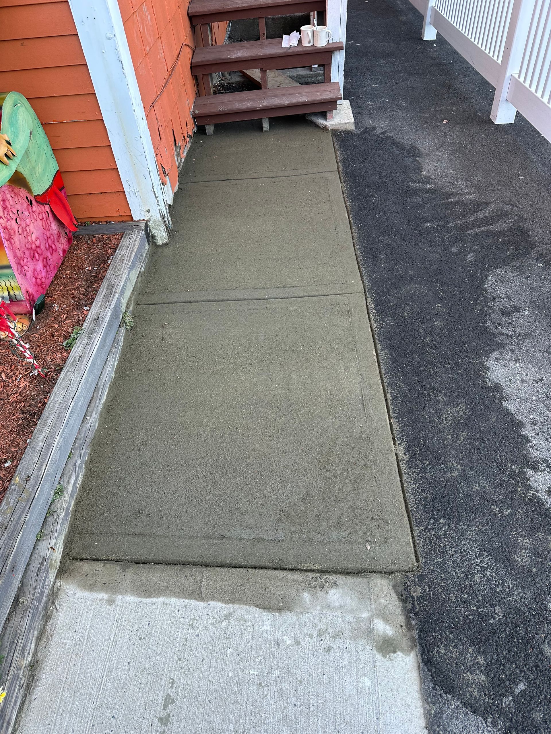 Narrow concrete walkway beside a red house, with steps ahead and asphalt driveway on the right.