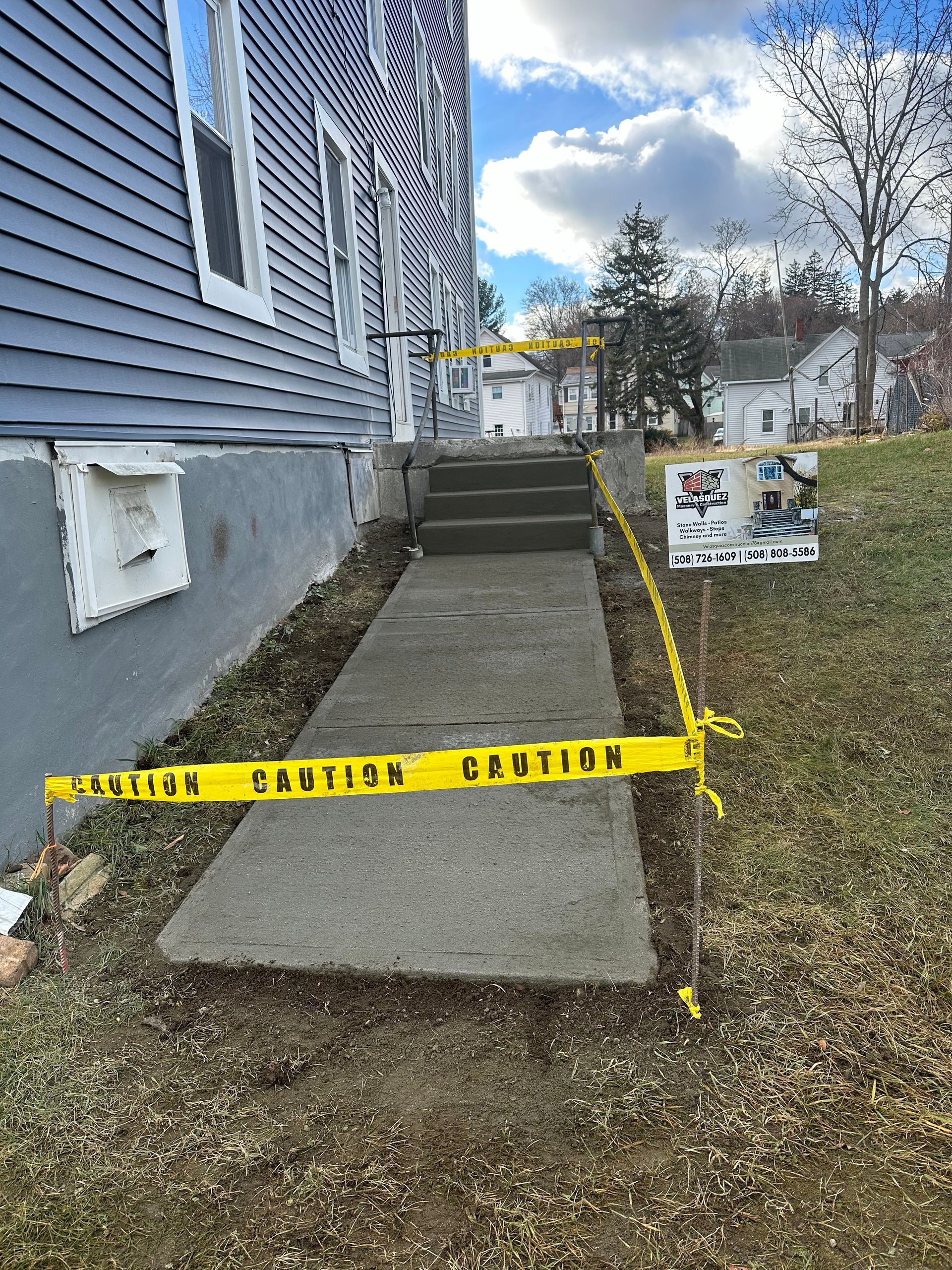 Yellow caution tape blocks a concrete walkway beside a blue house under construction.