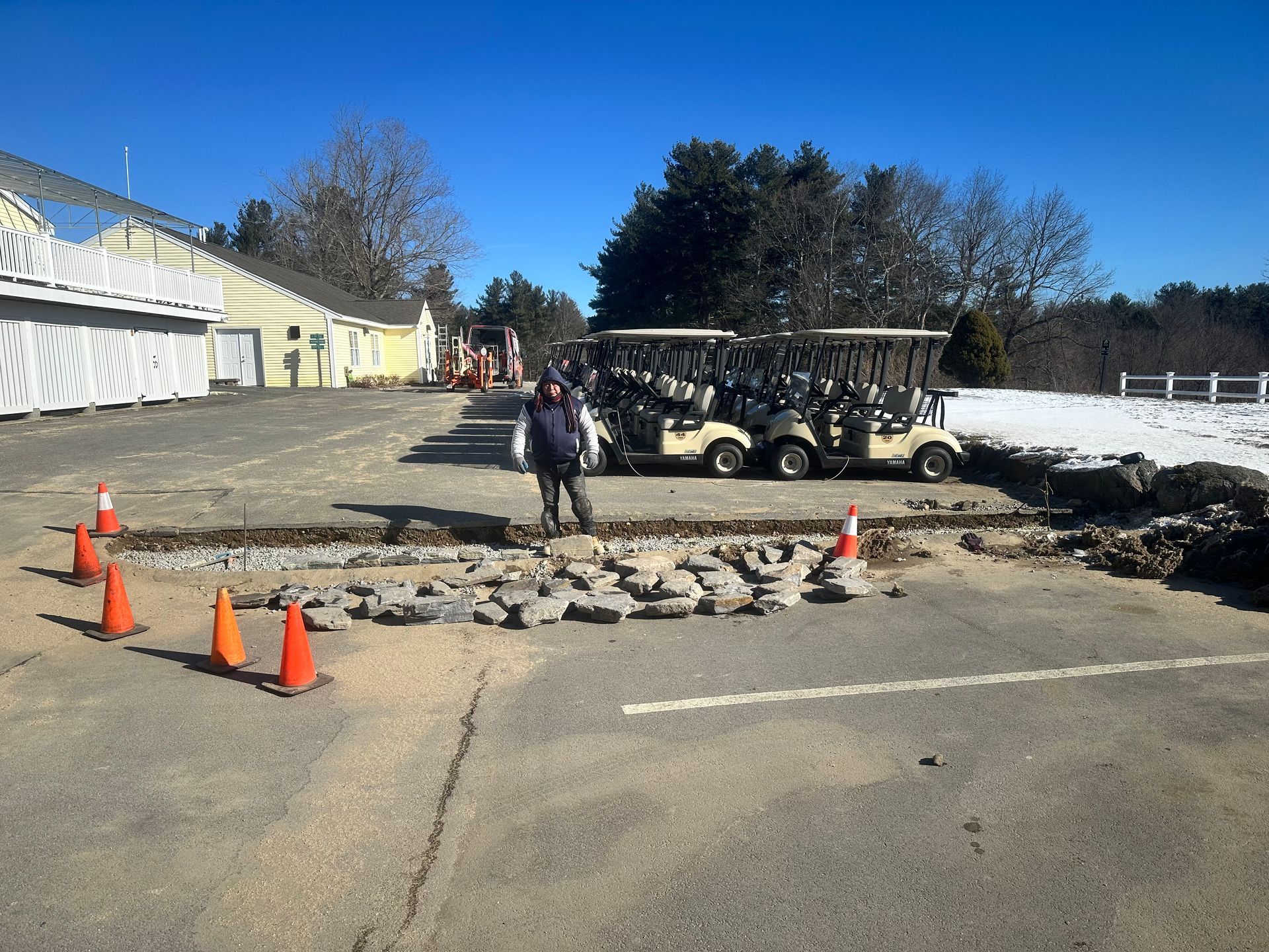 Workers repairing a pothole in a parking lot, with traffic cones and vehicles nearby.